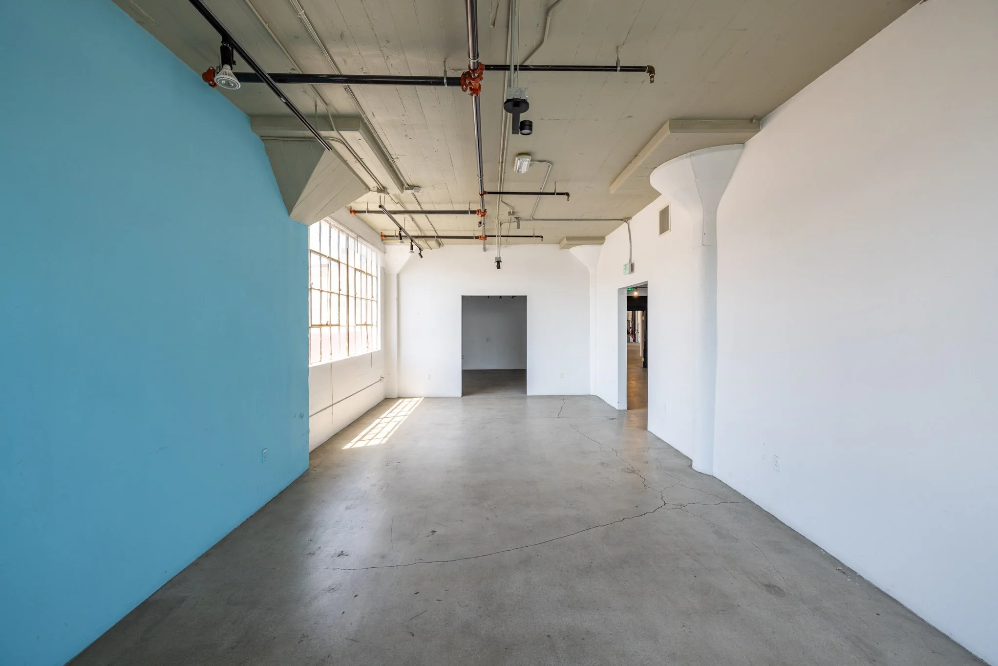 Interior of an empty room with white walls, a large window casting sunlight on the concrete floor, and an exposed ceiling with pipes and lighting fixtures.