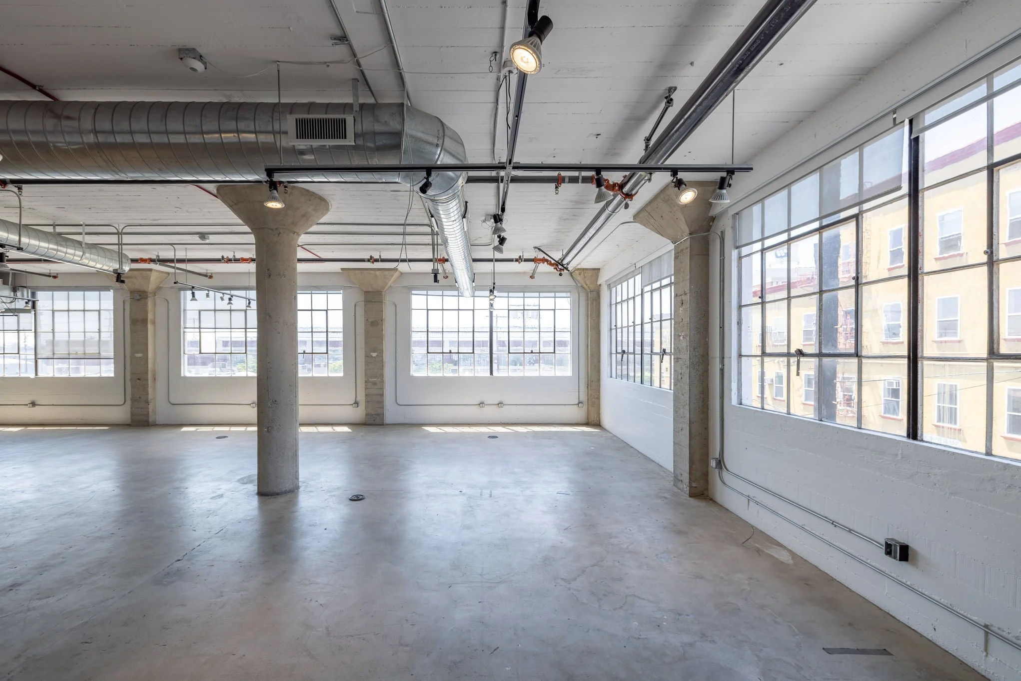 Empty industrial-style loft with large windows, concrete floor, exposed ducts, and support columns.