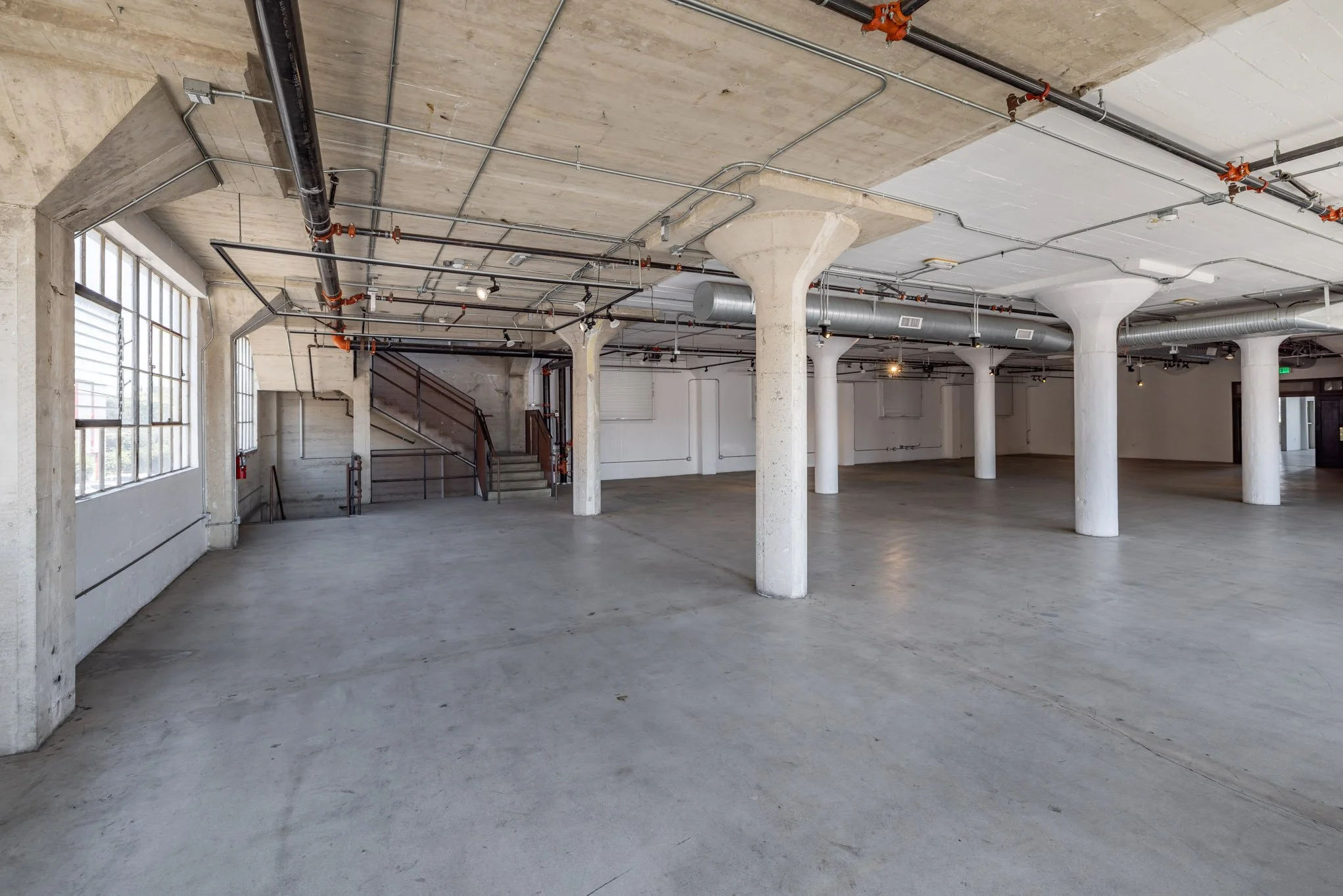 Empty industrial-style interior space with concrete floors, large windows, white columns, and exposed ceiling pipes.