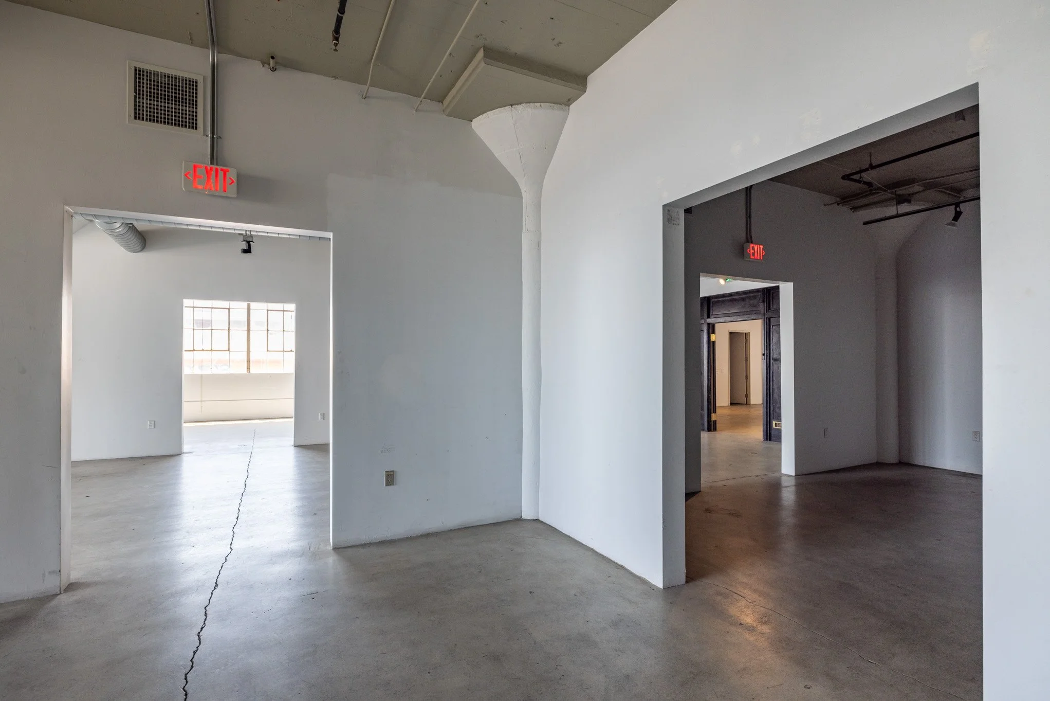 Interior of a modern building with white walls, concrete floors, exposed ceiling pipes, and multiple doorways leading to different rooms, some with visible exit signs.
