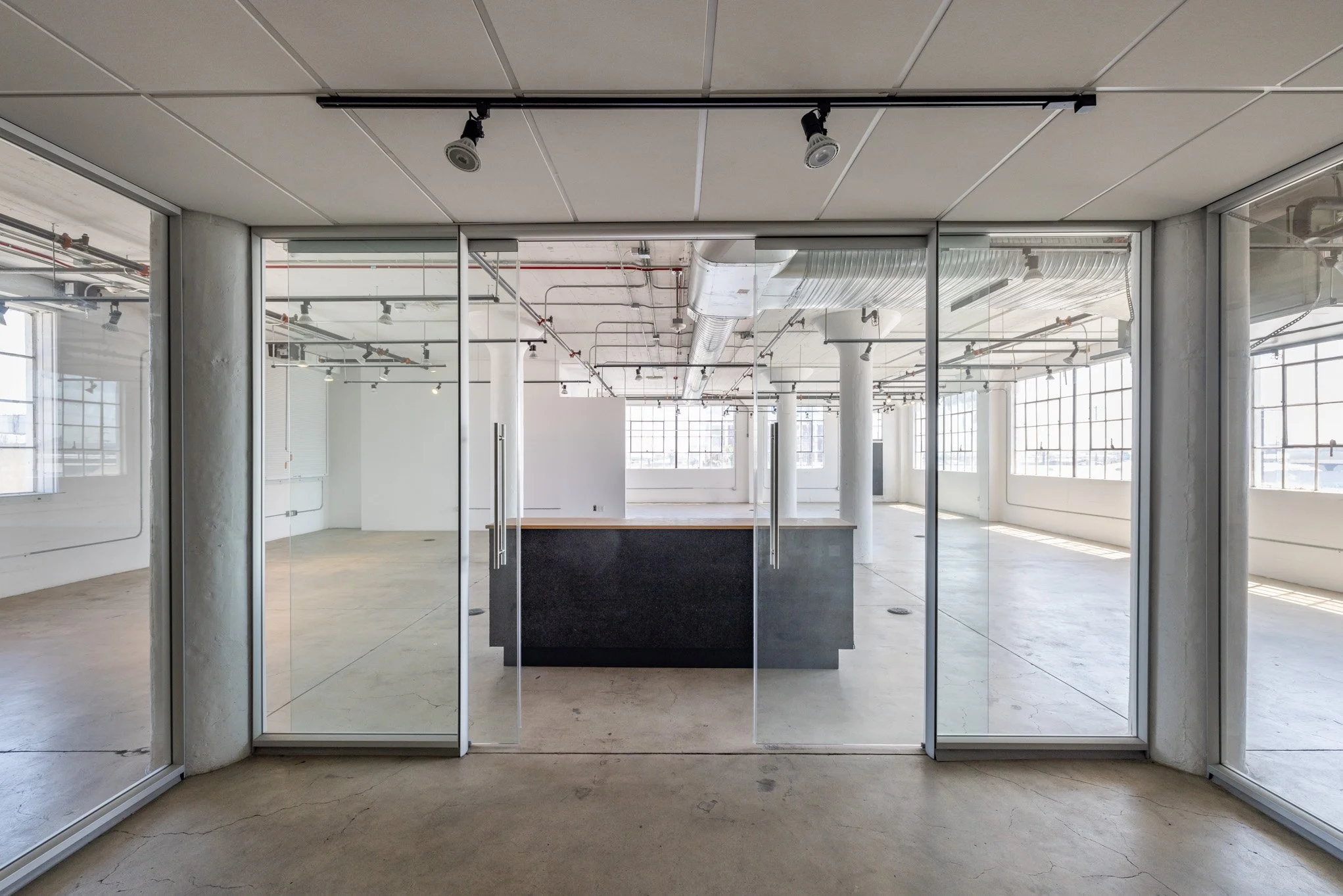 Empty commercial space with large windows, glass doors, concrete floors, and exposed ceiling pipes, seen from outside through glass door.