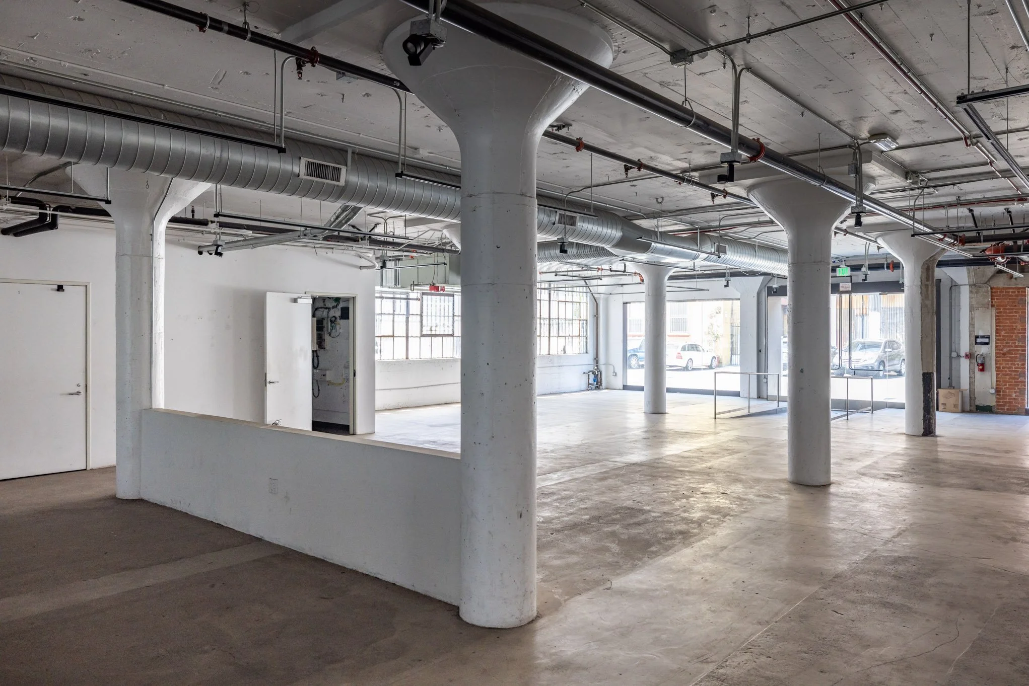 An empty industrial-style commercial space with concrete pillars and exposed ceiling pipes, large windows allowing natural light, and a mix of concrete and wood flooring.