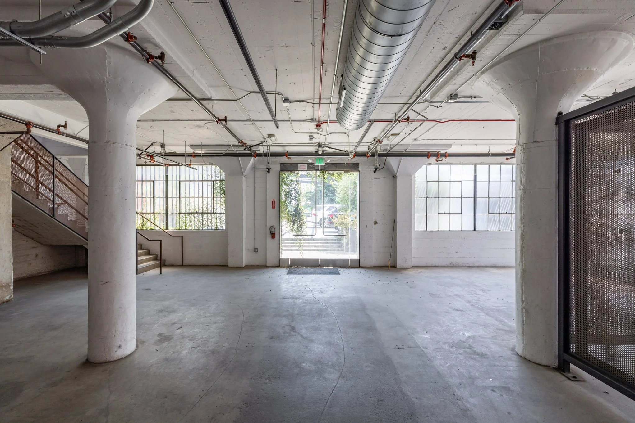 Empty industrial or commercial space with concrete floors, large windows, white walls, and exposed ductwork on the ceiling.