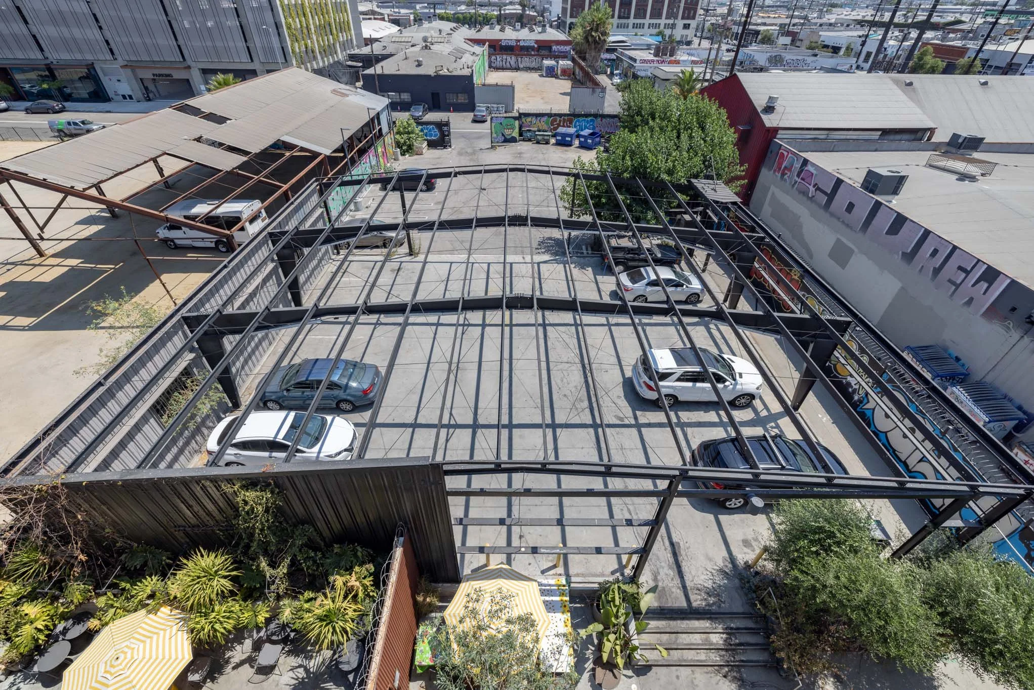 Empty parking lot bordered by a black metal framework, with a small outdoor seating area with yellow and white umbrellas and greenery.