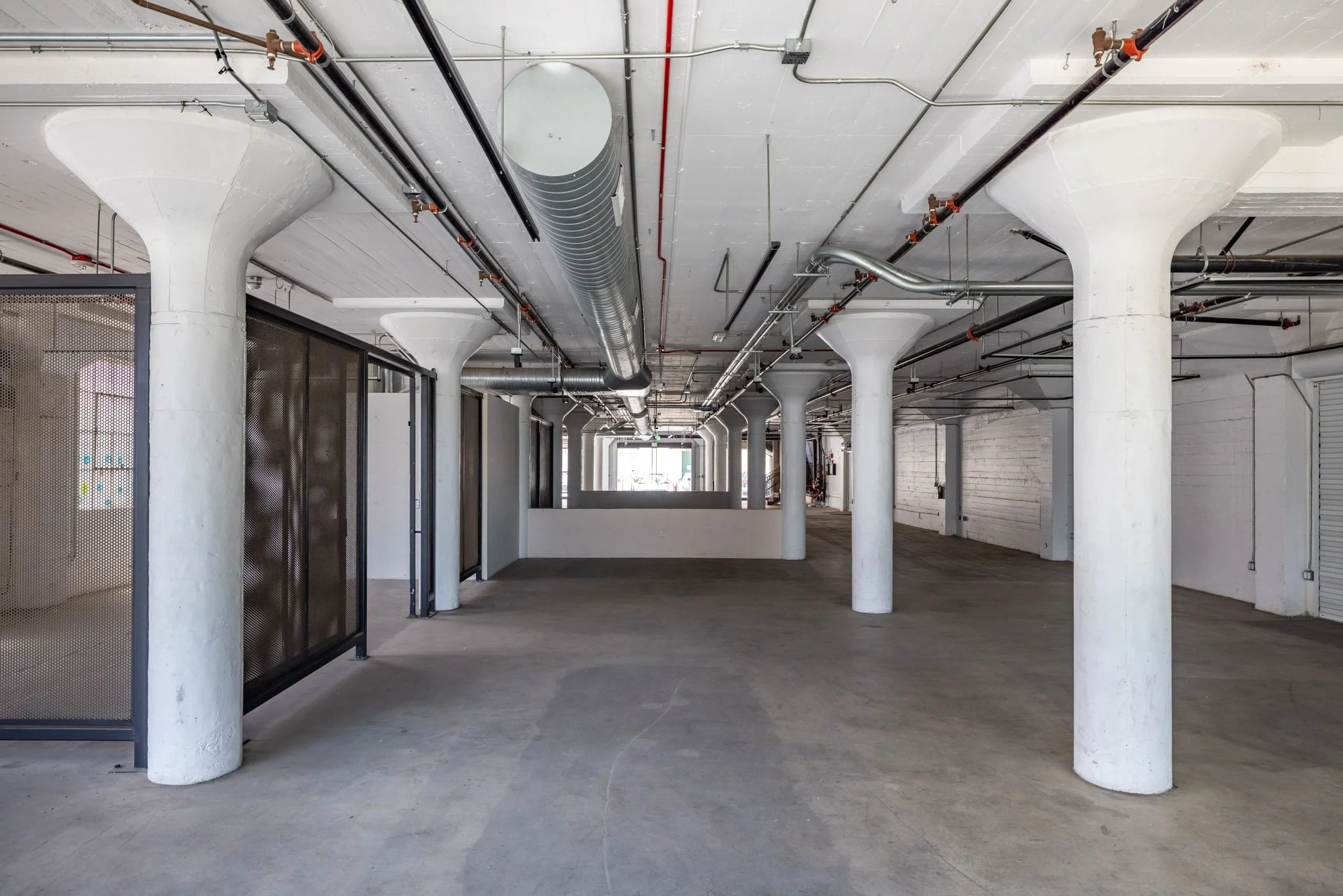 Empty industrial or commercial space with large white concrete pillars, exposed ceiling pipes, and a concrete floor.