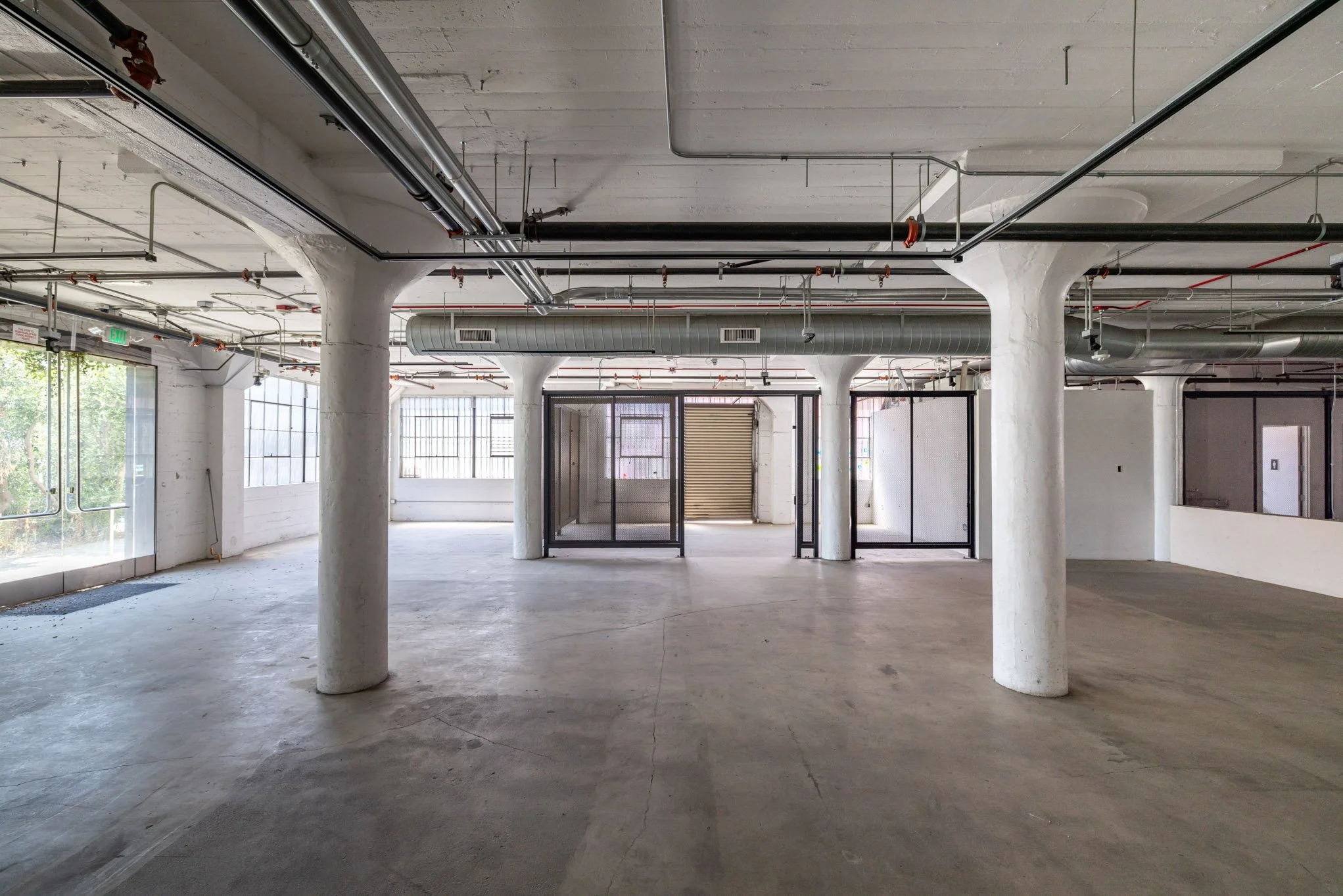 Empty industrial-style interior space with white concrete pillars, large windows, exposed ductwork, and glass partition walls.