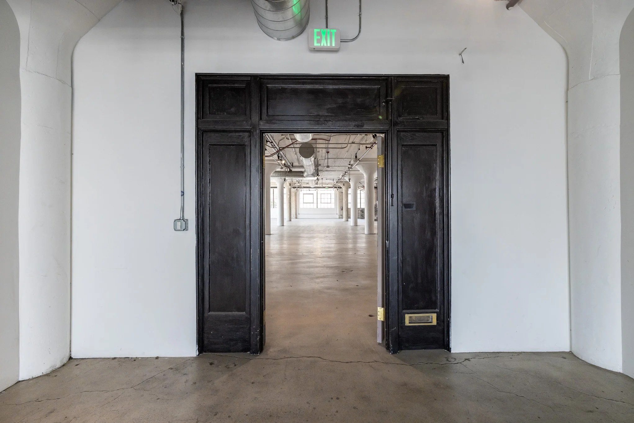 Open black door leading into a bright, spacious industrial-style interior with white columns and large windows.