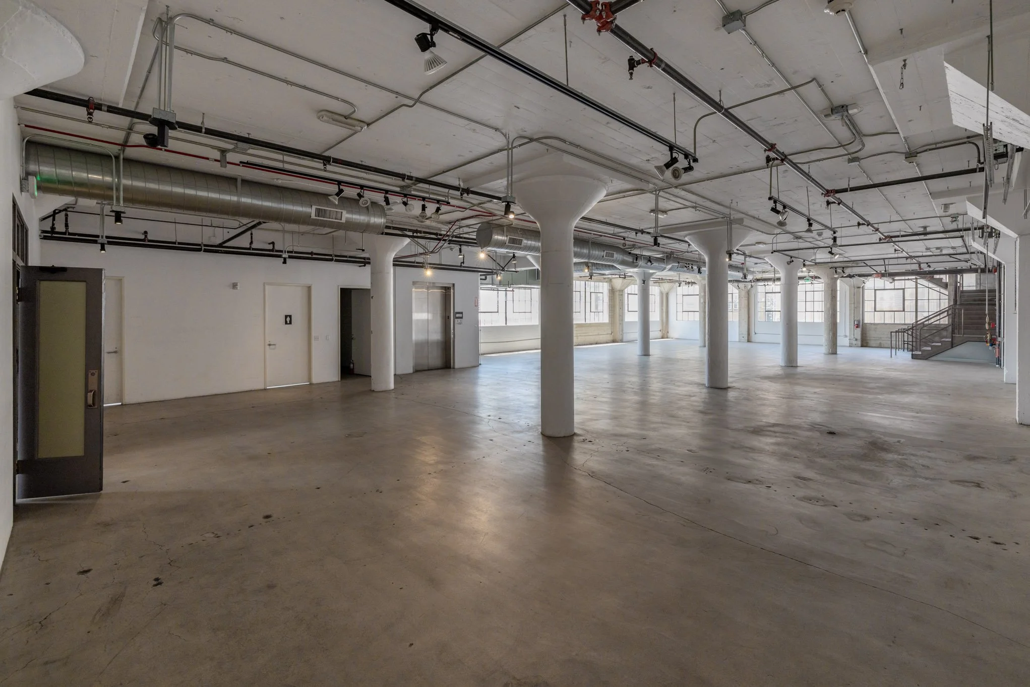 Empty industrial-style interior space with concrete floors, white walls, exposed ductwork, and large windows, featuring an elevator and staircase in the background.