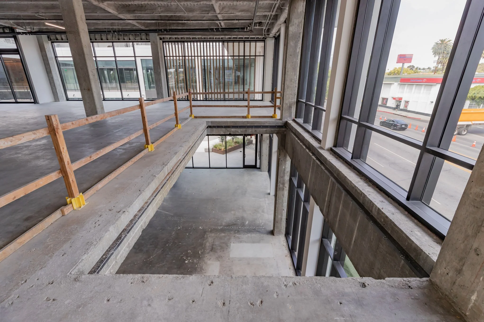 Interior of a multi-story building under construction with unfinished concrete floors, large windows, and wooden safety barriers installed along the edge of the upper floor.