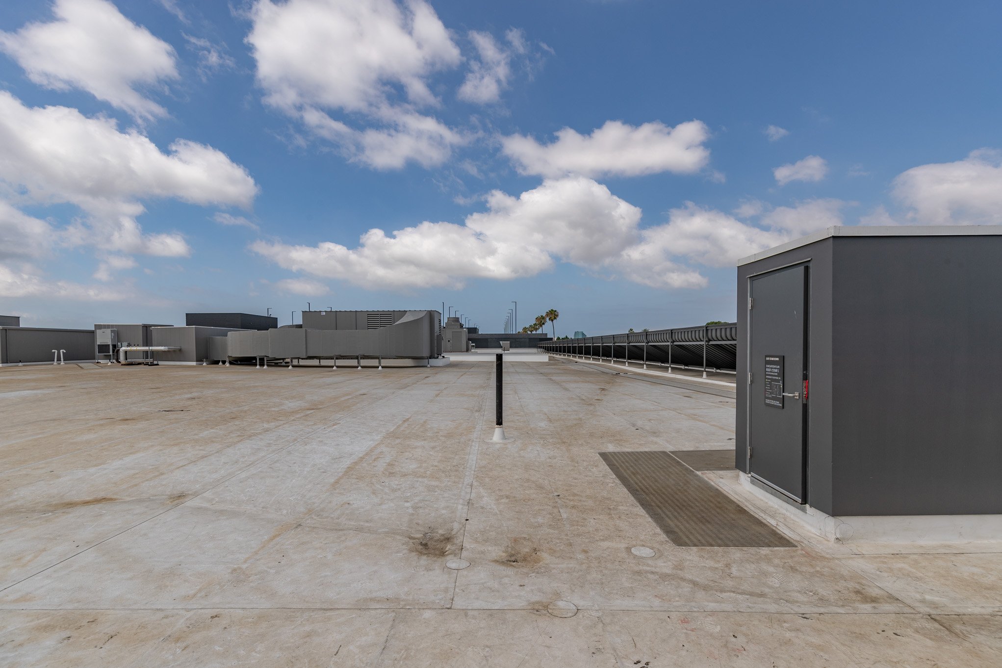 Empty rooftop with a clear blue sky, scattered white clouds, and rooftop equipment including vent systems and a gray electrical enclosure.