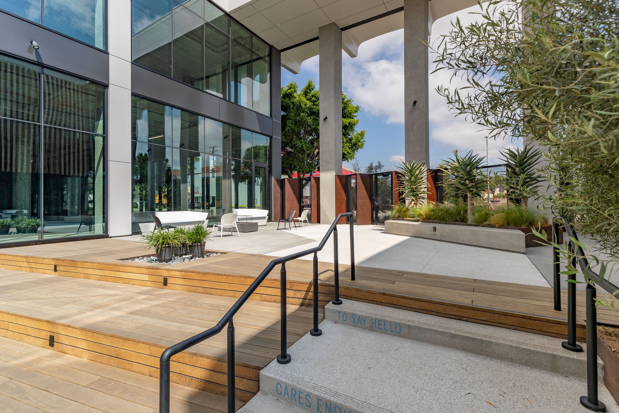 Modern outdoor patio with glass building, wooden steps, potted plants, and seating area, surrounded by trees and greenery.