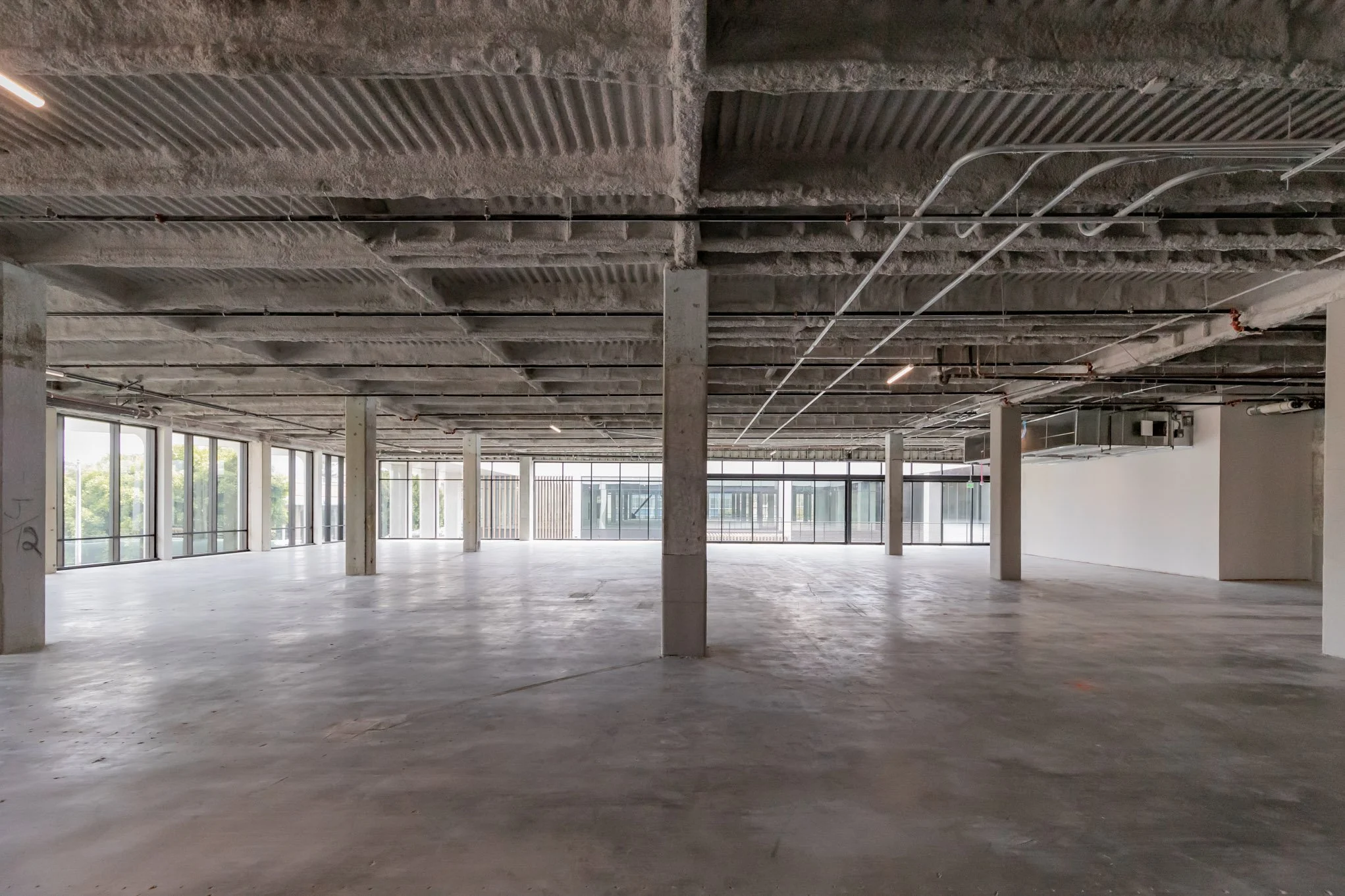 Interior view of an unfinished, spacious commercial building with exposed concrete floors, ceiling, and support columns. Large windows along the walls let in natural light.