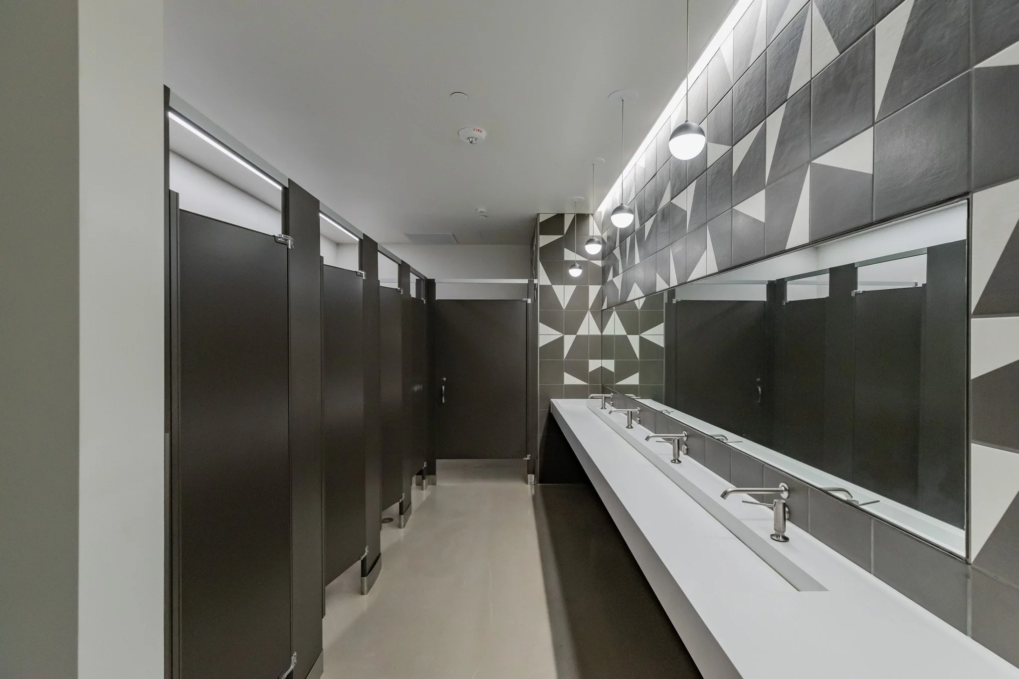 Public restroom with black stalls and a long white sink with multiple faucets, gray geometric tile wall, and circular ceiling lights.