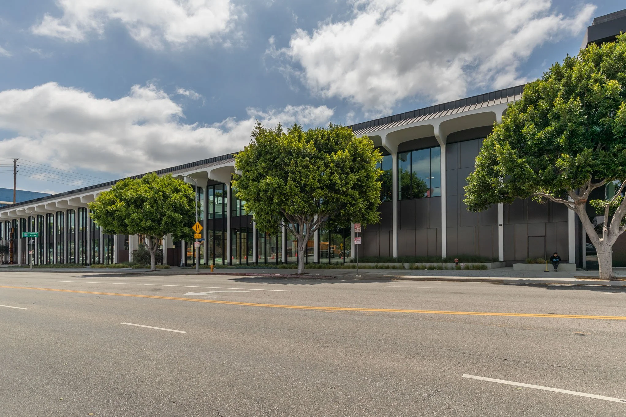 Modern building with large glass windows, surrounded by green trees, on a city street under partly cloudy sky.