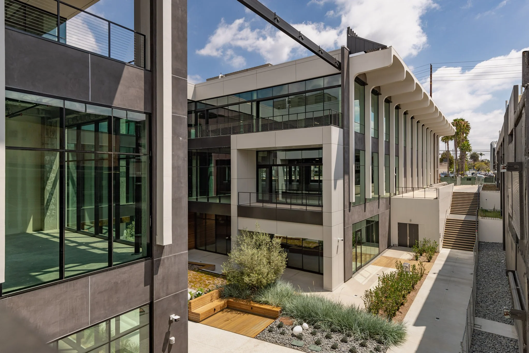 Modern multi-story building with large glass windows, white and black exterior walls, and outdoor landscaping featuring shrubs, trees, and benches, under a partly cloudy sky.