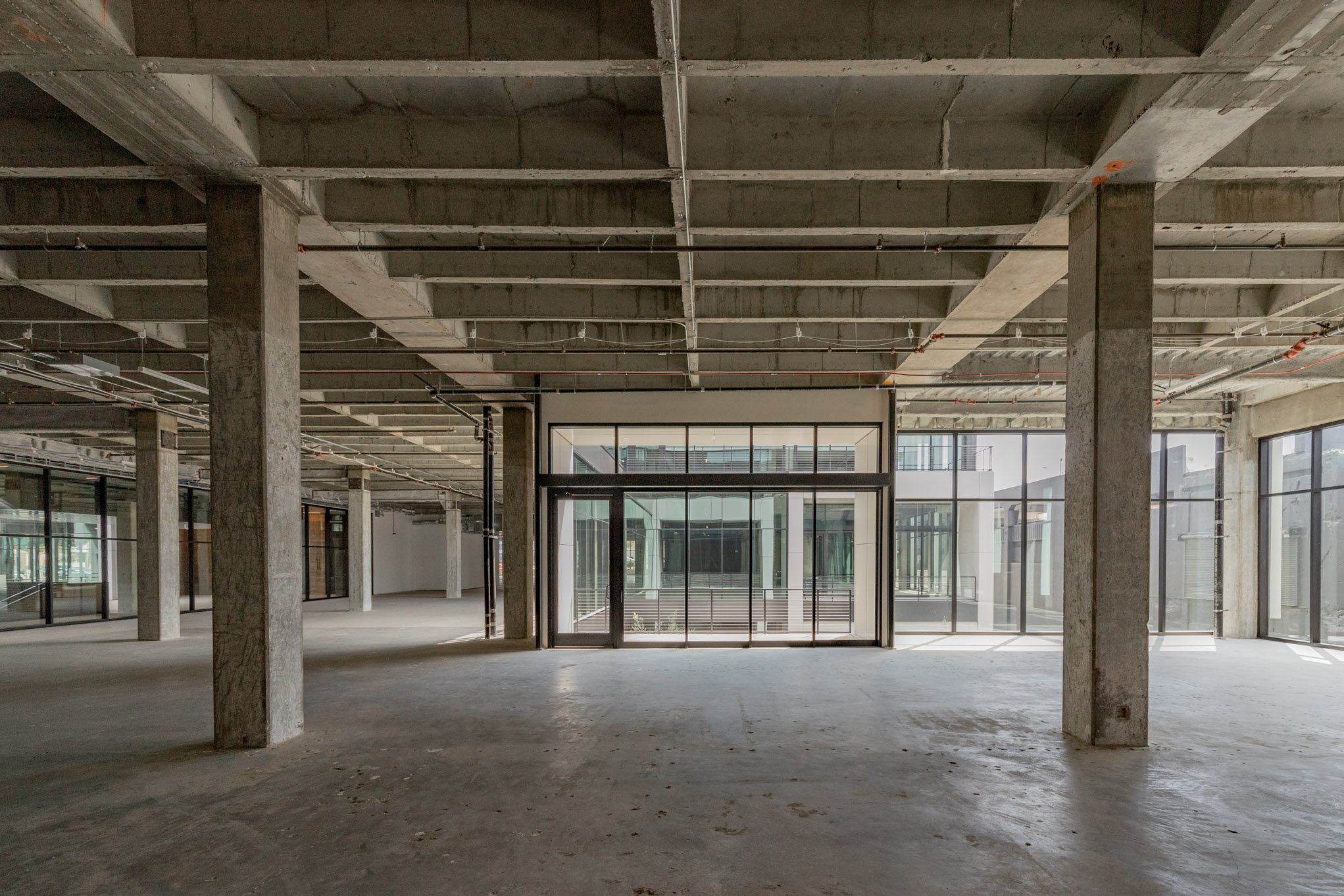 An unfinished commercial interior space with exposed concrete columns and floors, metal ceiling structure, and large glass doors and windows letting in natural light.