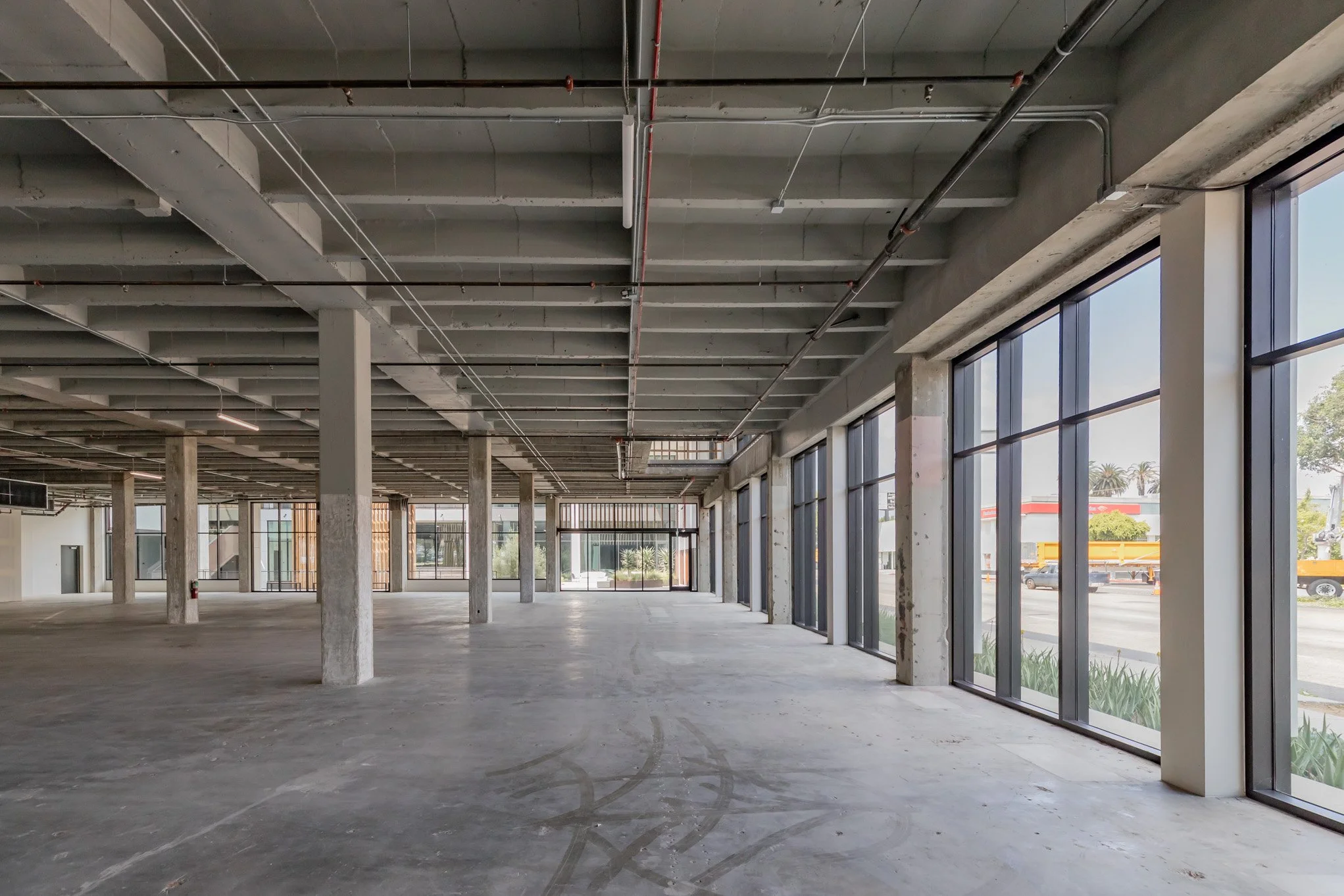 Empty commercial space with large floor area, concrete columns, unfinished ceiling, and floor-to-ceiling glass windows along one wall showing a view of the street and parking area.