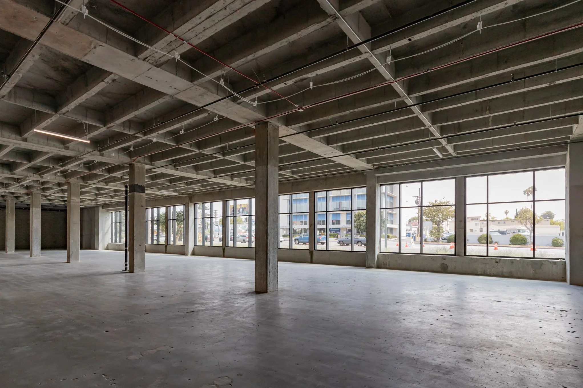 Empty commercial space with large windows, concrete floors, and exposed ceiling beams.