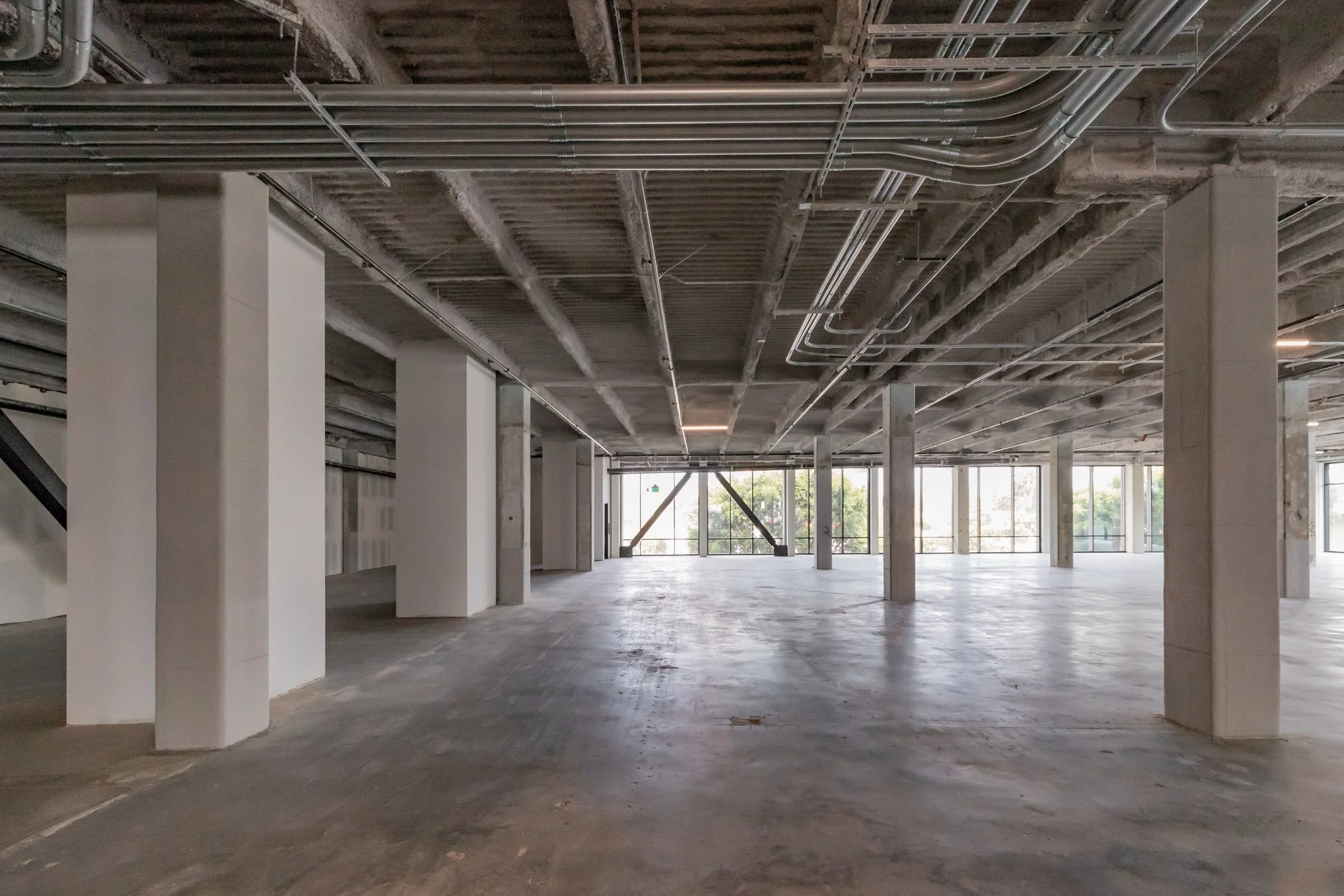Large unfinished interior space with concrete floors, white walls, exposed ceiling with pipes, and large windows letting in natural light.