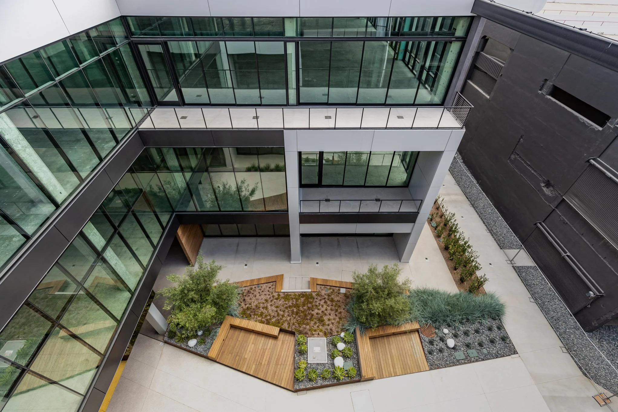An aerial view of a modern building courtyard with plants, trees, and benches, surrounded by glass windows and dark-colored walls.