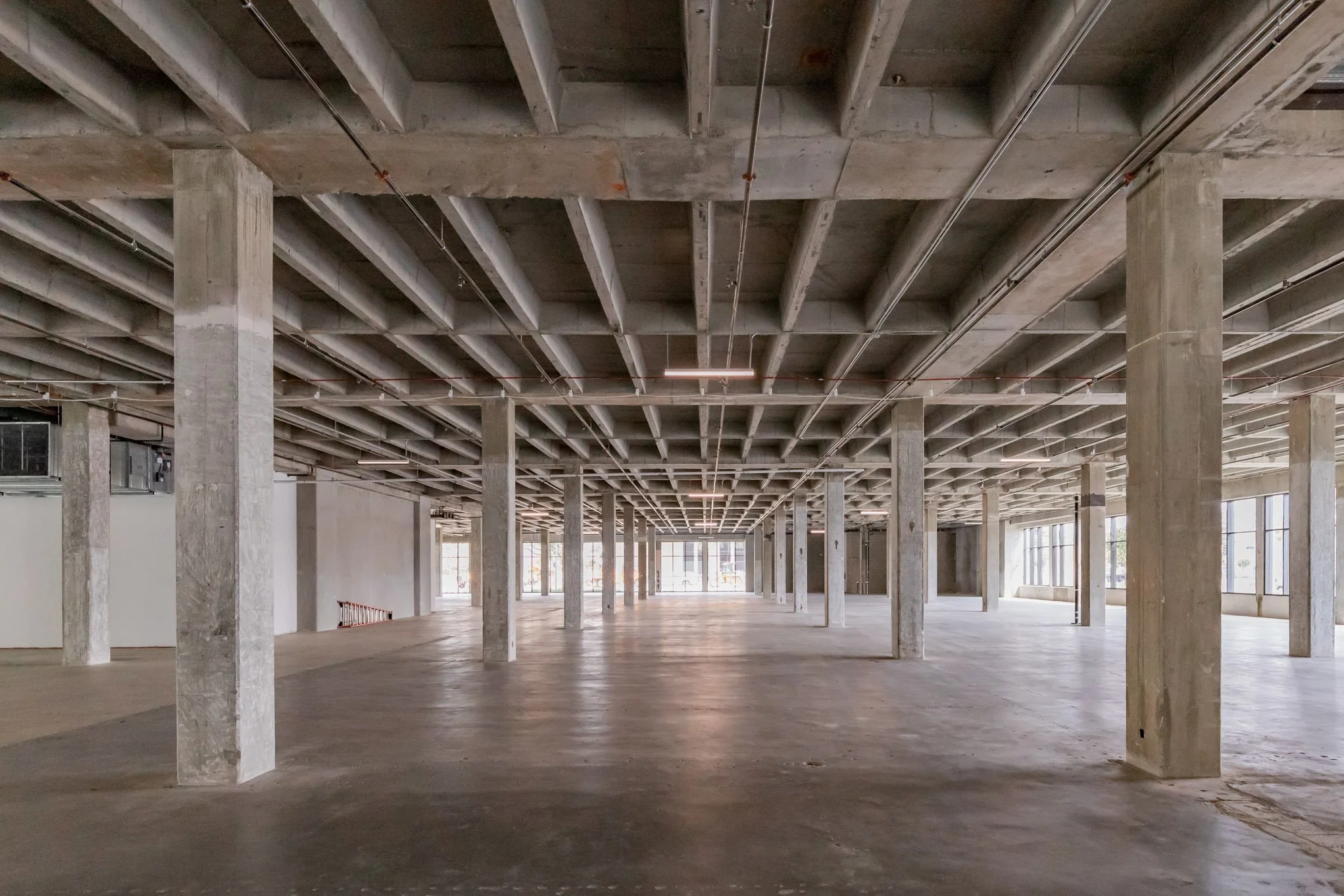Empty commercial space with concrete floors, exposed concrete columns, and an unfinished ceiling with visible beams and piping, with large windows letting in natural light.