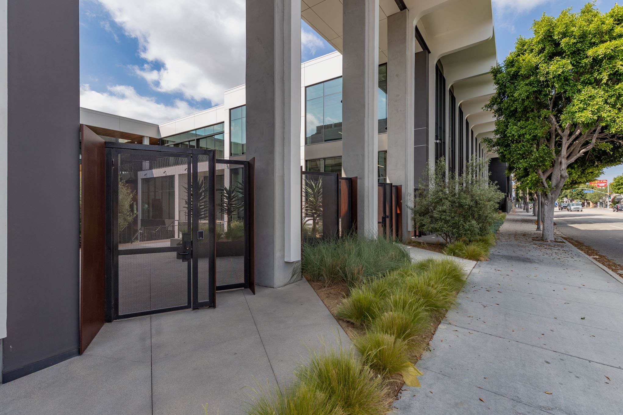Modern building with glass windows, concrete columns, a sidewalk, green trees, and landscaped plants