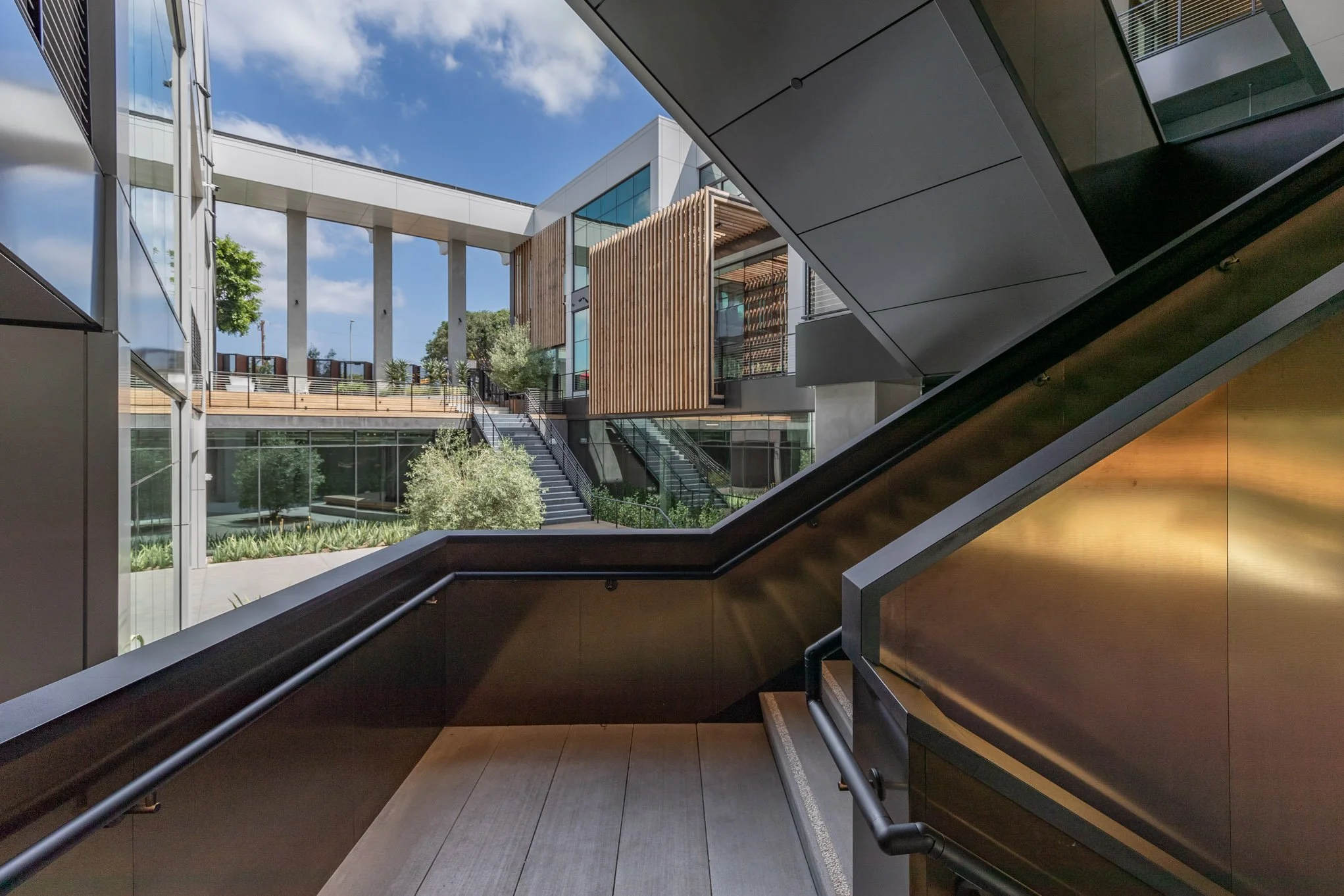 View of an outdoor multi-level modern building courtyard with stairs, glass walls, wooden accents, greenery, and a blue sky with white clouds.