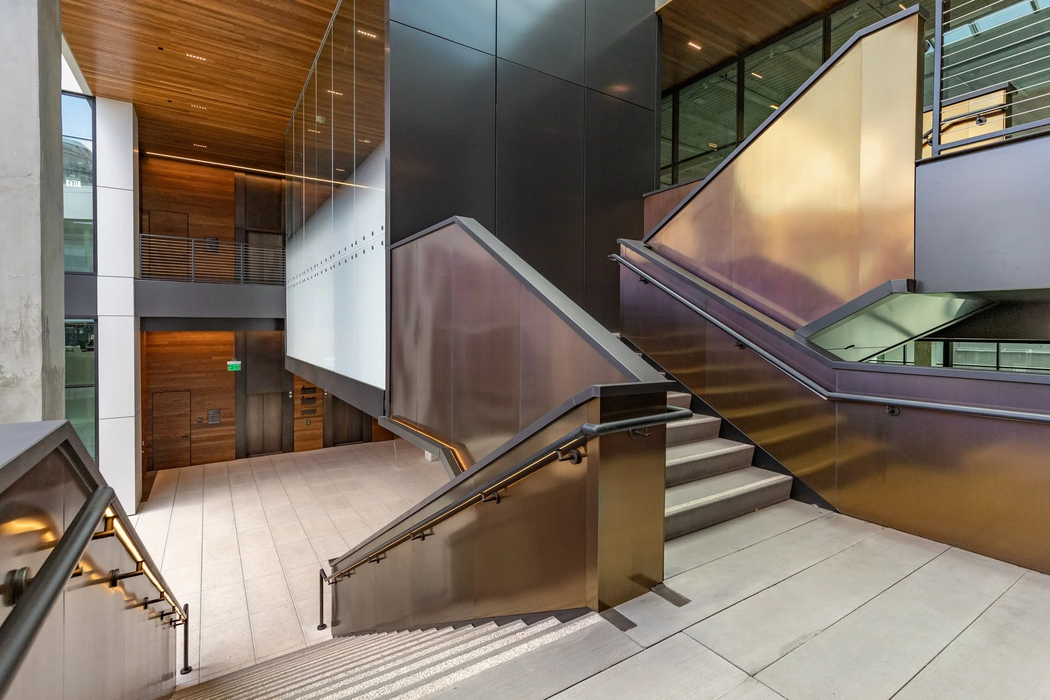 Modern interior with staircase, glass railings, wood, black and metallic surfaces, and natural light.