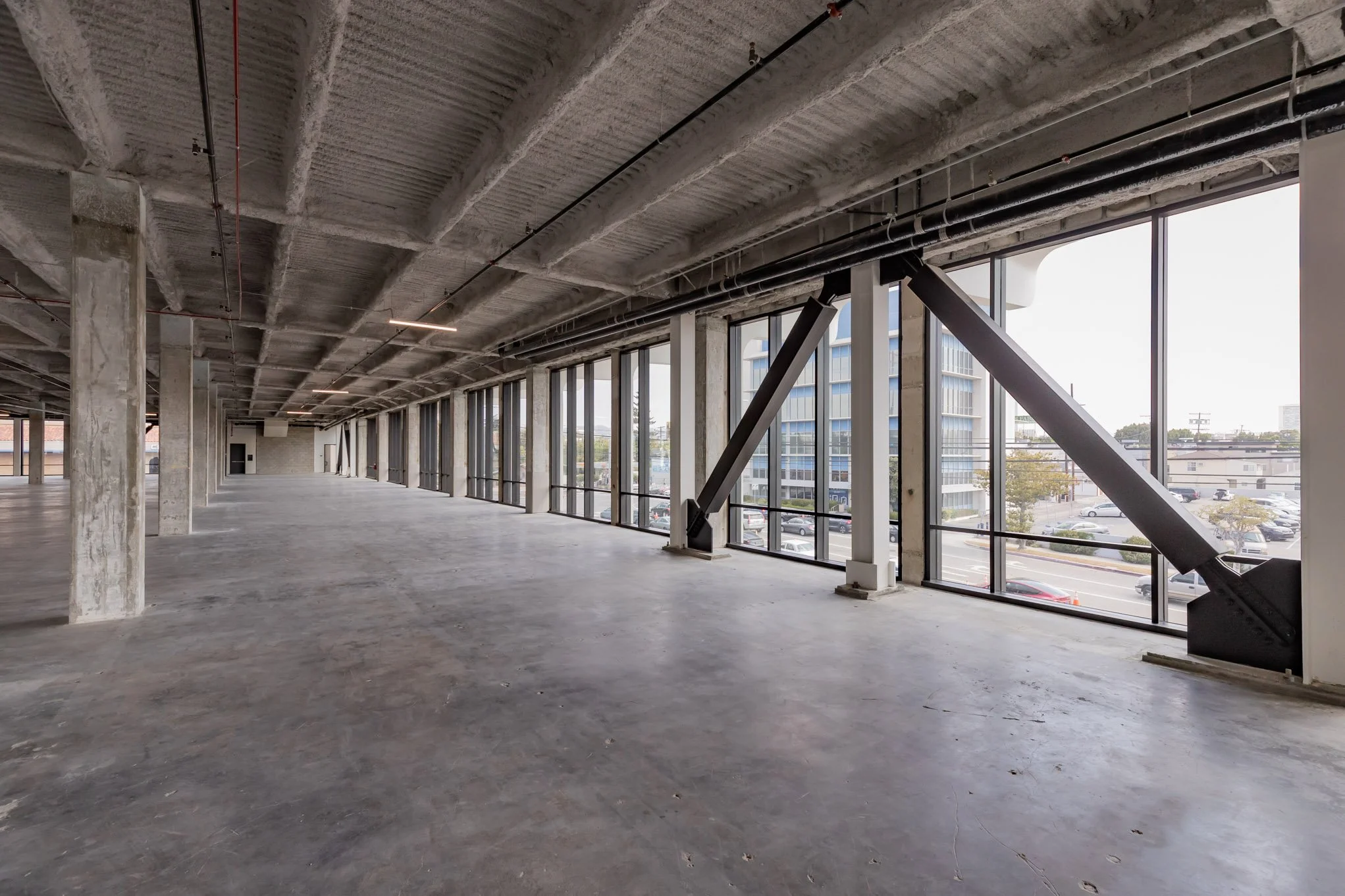 Empty commercial building with concrete floors, exposed concrete pillars, unfinished ceiling, and large floor-to-ceiling windows showing city view.