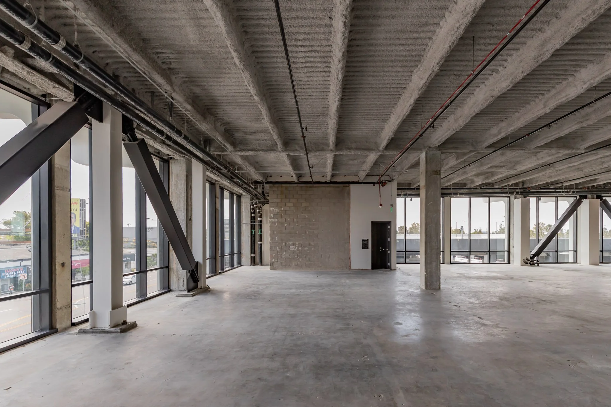 Empty commercial space under construction with large windows, concrete floors, and concrete ceiling with exposed beams.