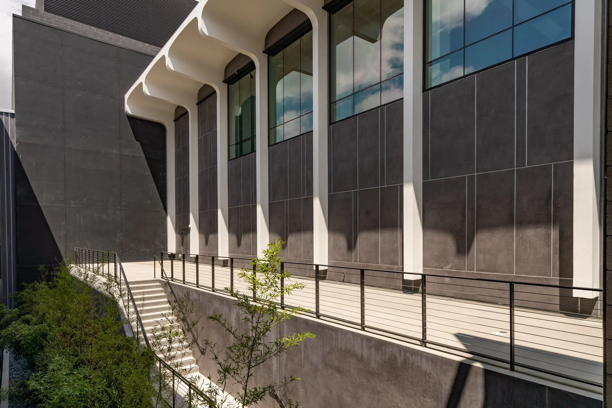 Modern building exterior with large glass windows, white structural supports, black metal railing, and stairs leading up to a balcony, with some greenery and a cloudy sky reflection.