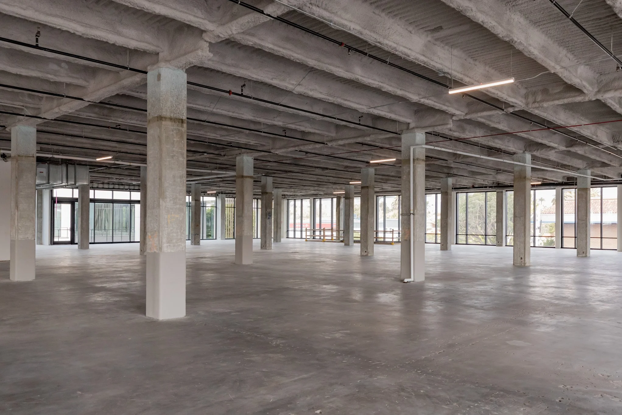 Empty, unfinished commercial space with concrete floors, concrete support columns, large windows, and exposed ceiling pipes and wiring.