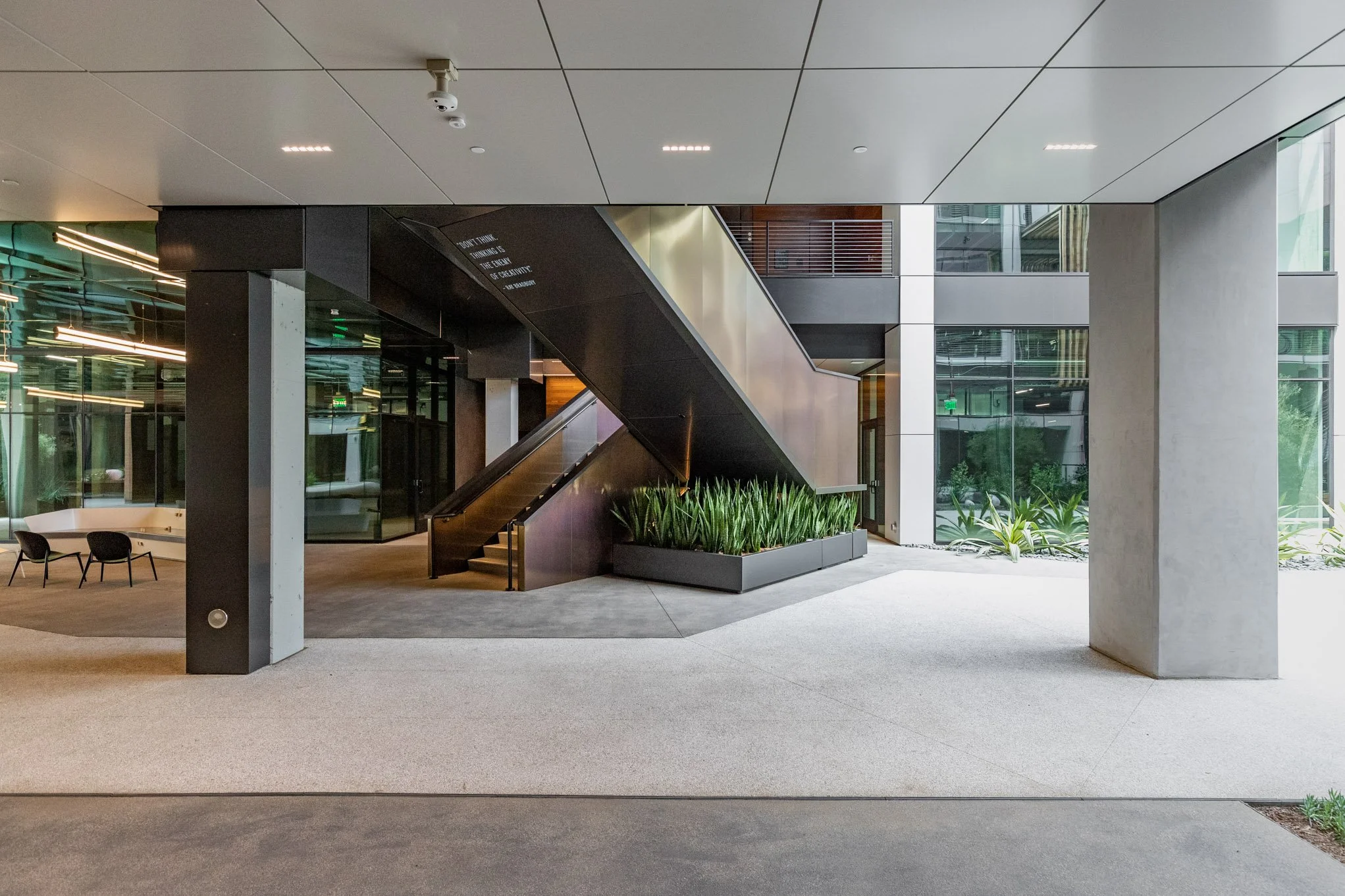 Modern office building lobby with staircase, large glass windows, indoor plants, and seating area