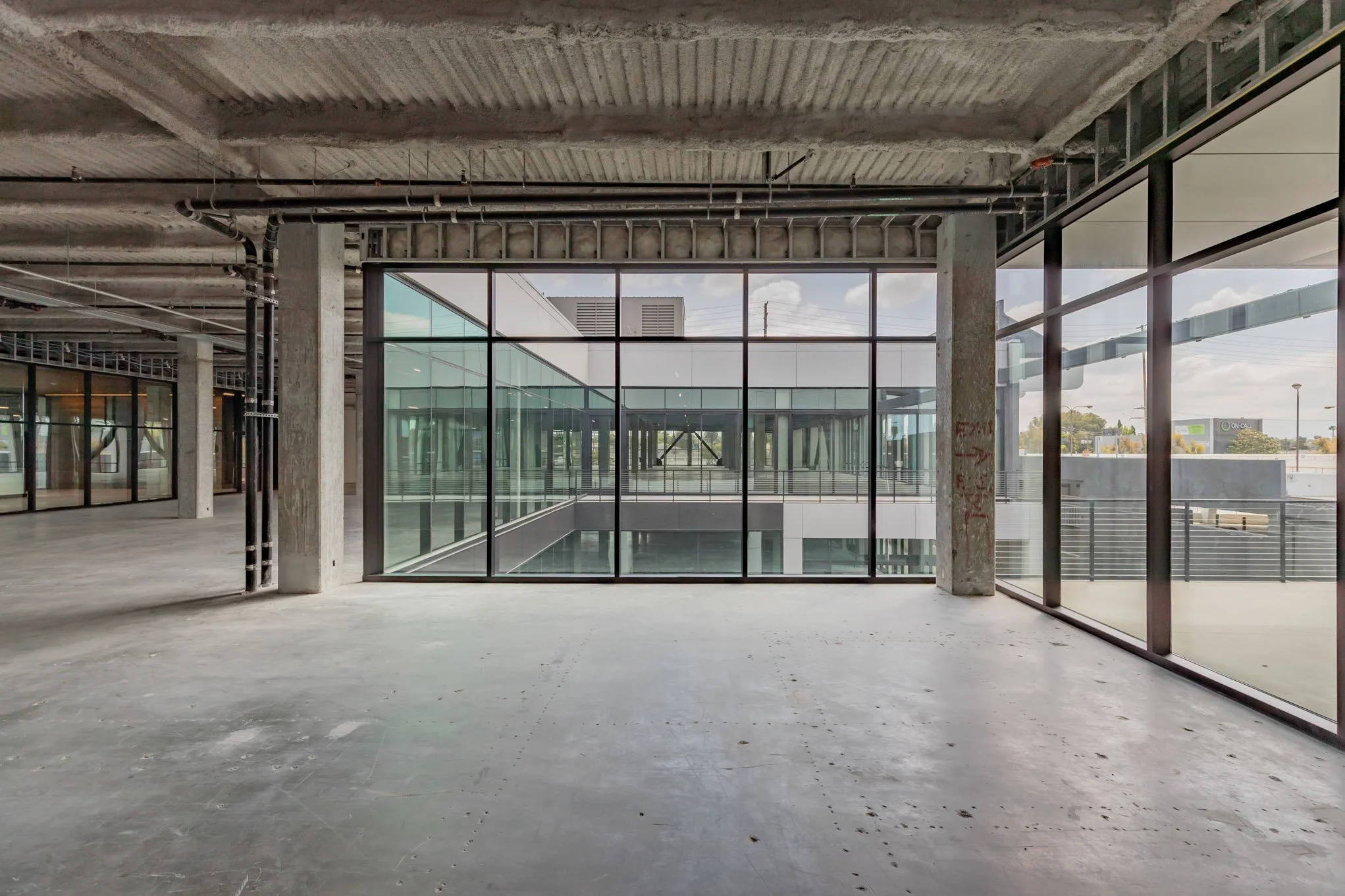 Empty interior space in a modern building under construction, with concrete floors, unfinished walls, and large glass windows showing various parts of the exterior environment.