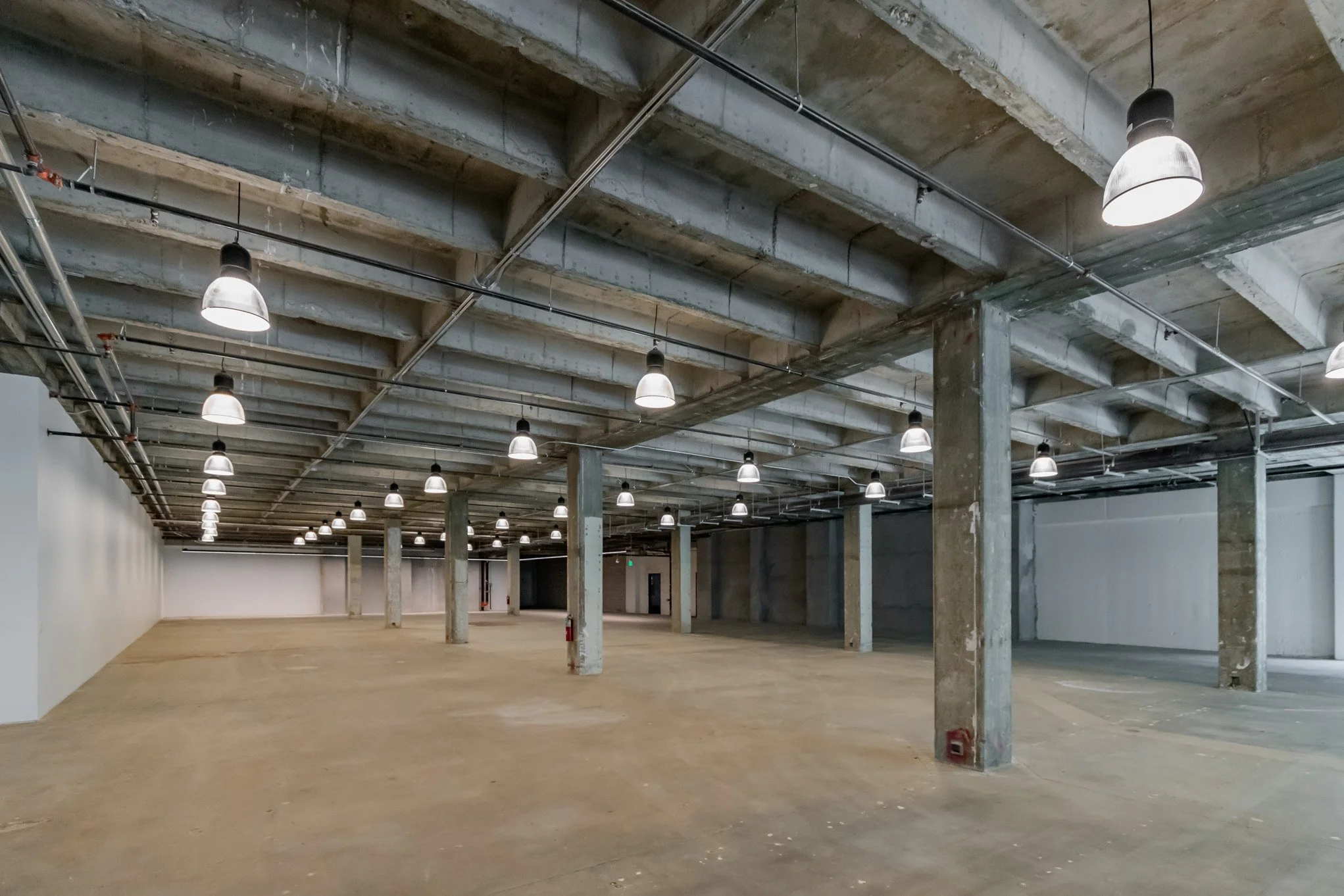 Empty industrial or warehouse space with concrete columns, ceiling beams, and hanging industrial lights.