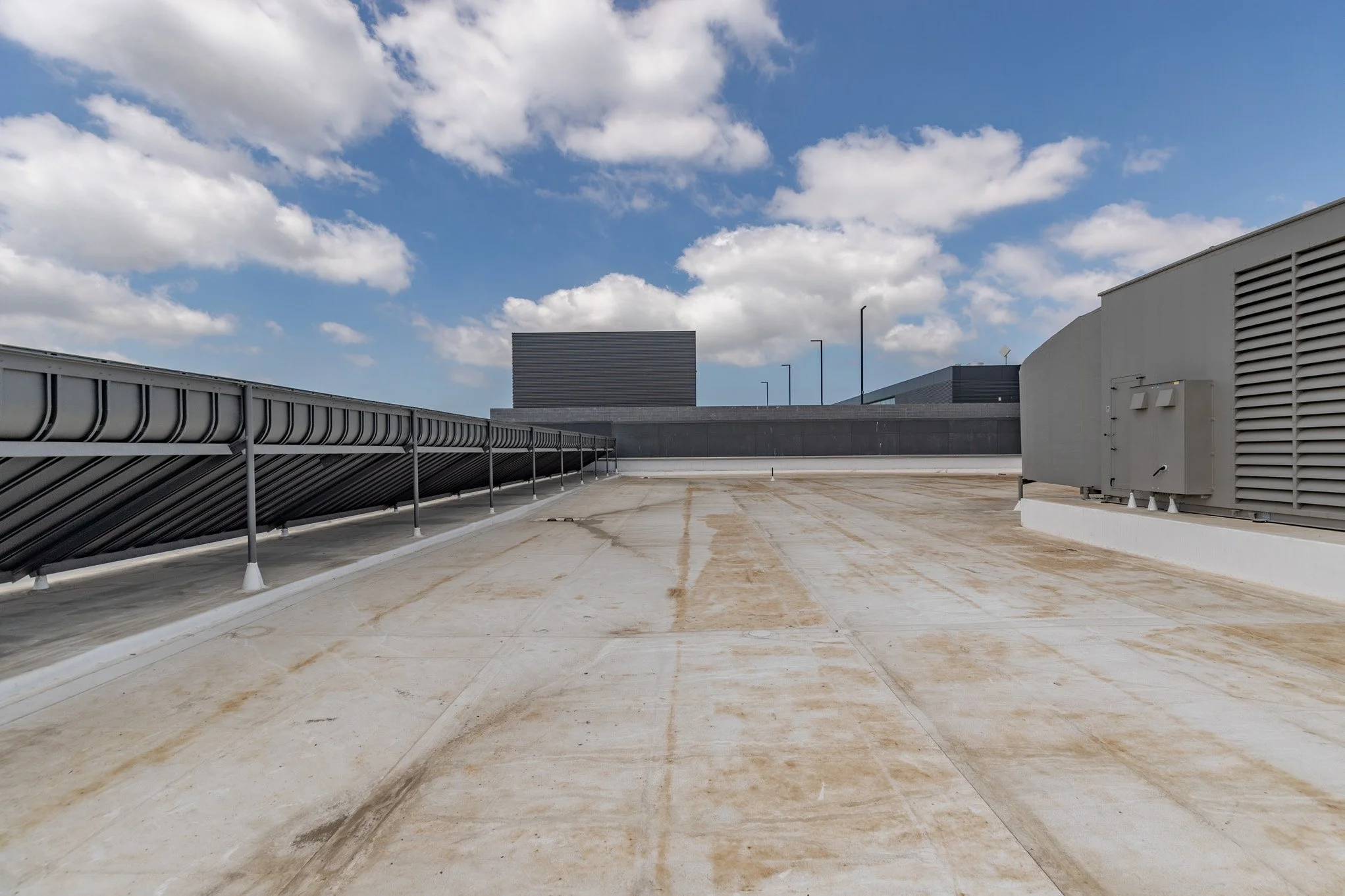 Rooftop with HVAC equipment, metal ventilation ducts, and a wall in the background under partly cloudy sky.
