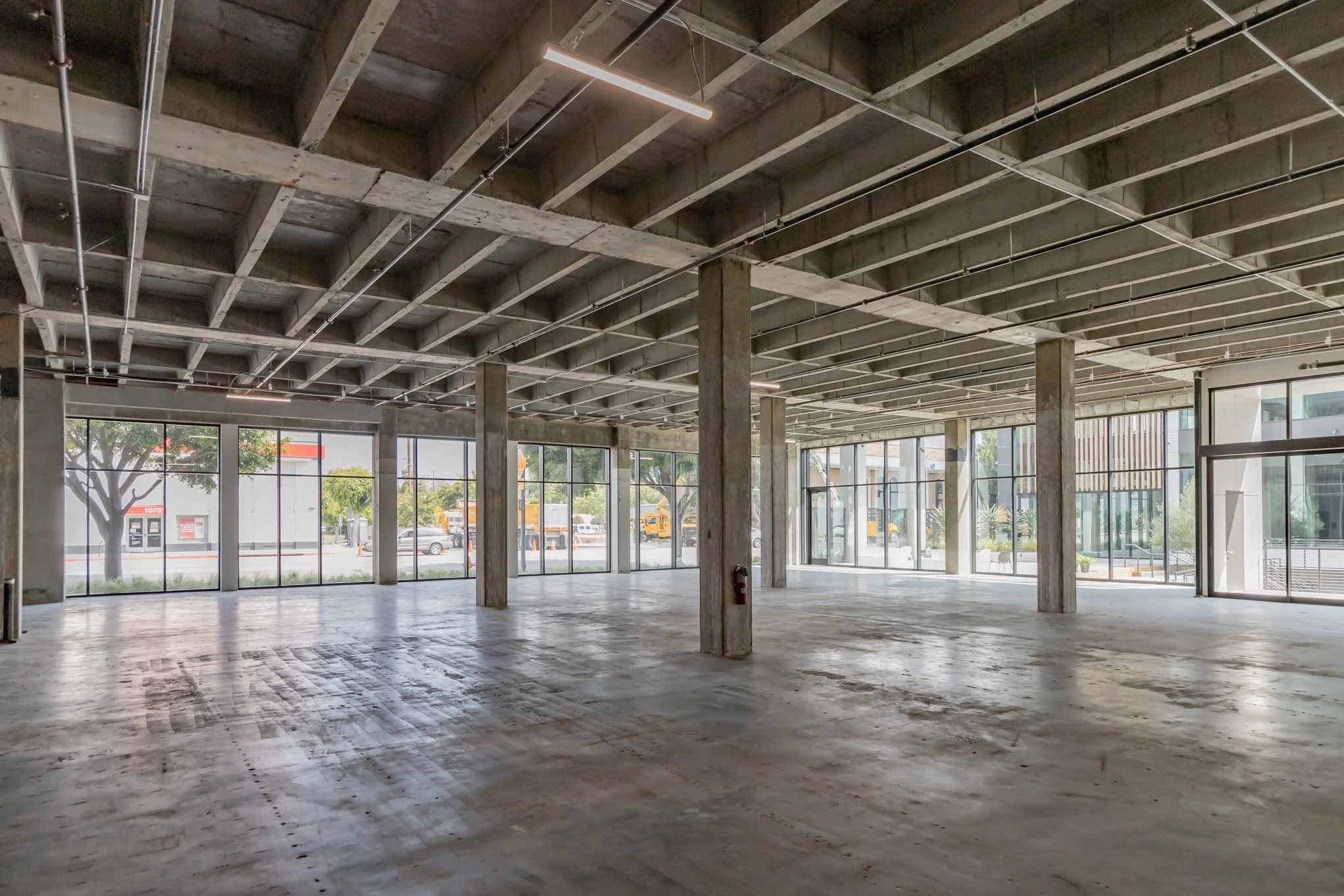 Empty commercial space with concrete floors, large glass windows, and exposed ceiling with lighting fixtures.