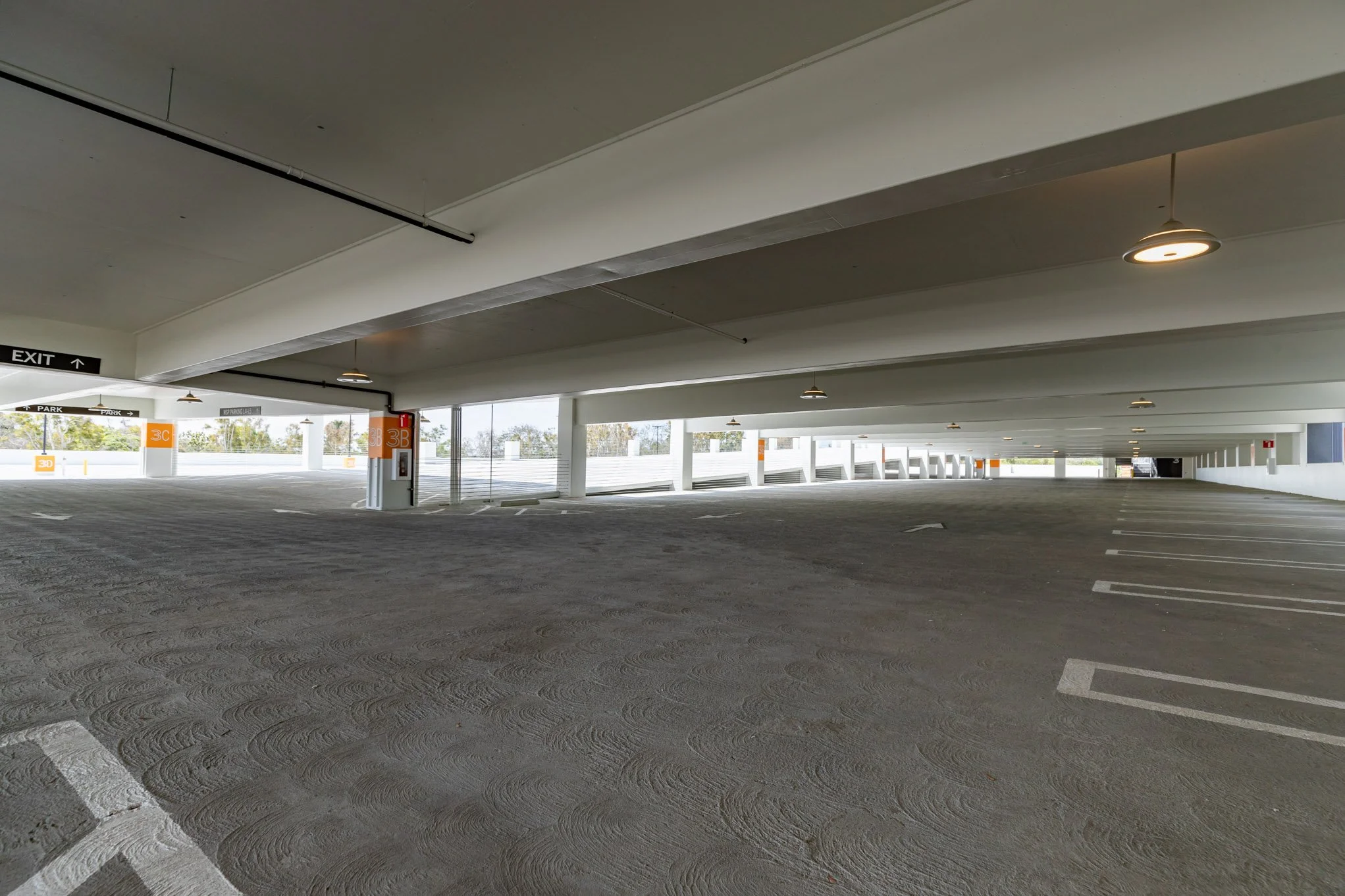 Empty multi-level parking garage with parking spaces and support columns, exit sign, and natural light from open sides.