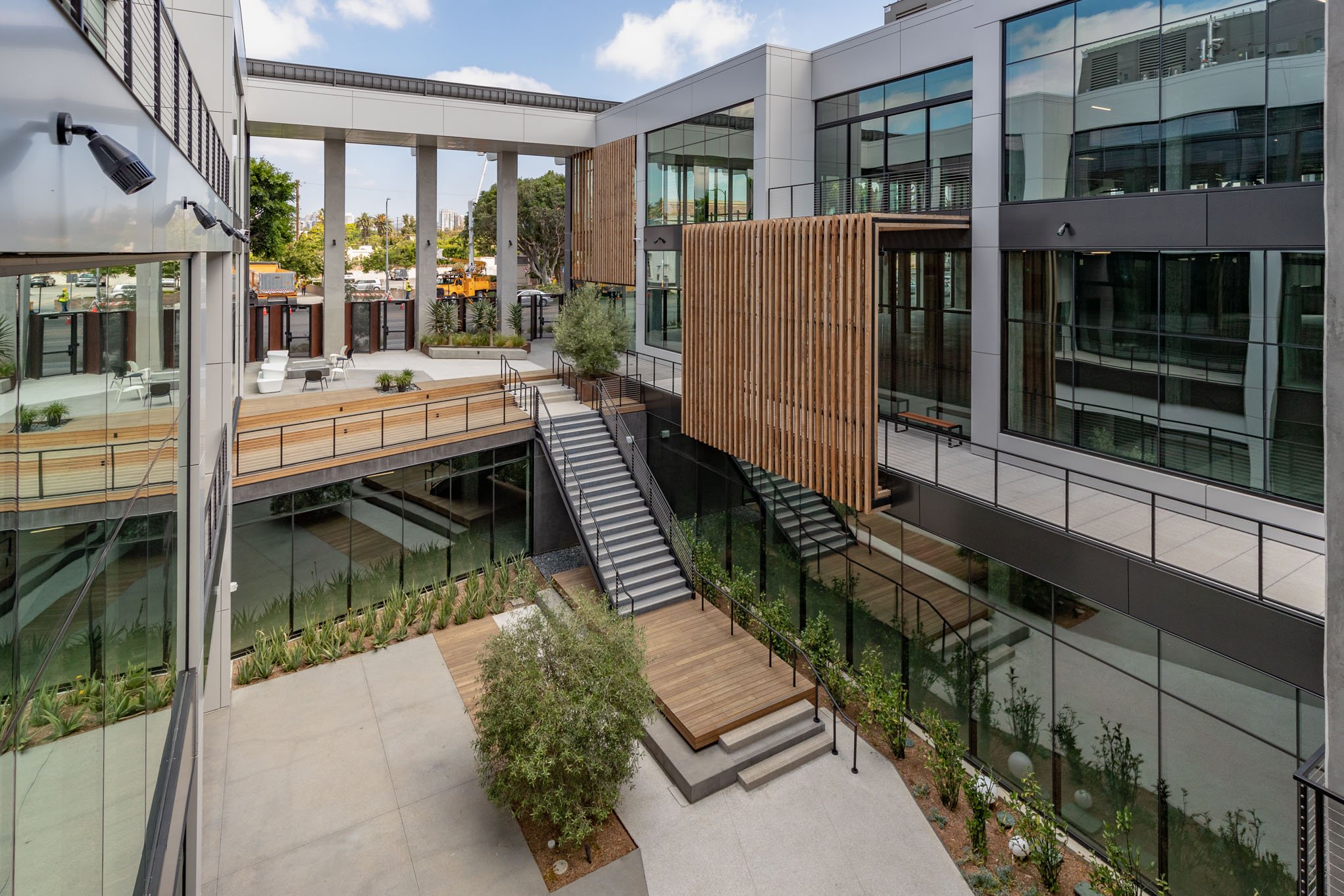 Modern multi-story building with glass exterior, outdoor staircases, wooden accents, and landscaped courtyard with trees and plants.