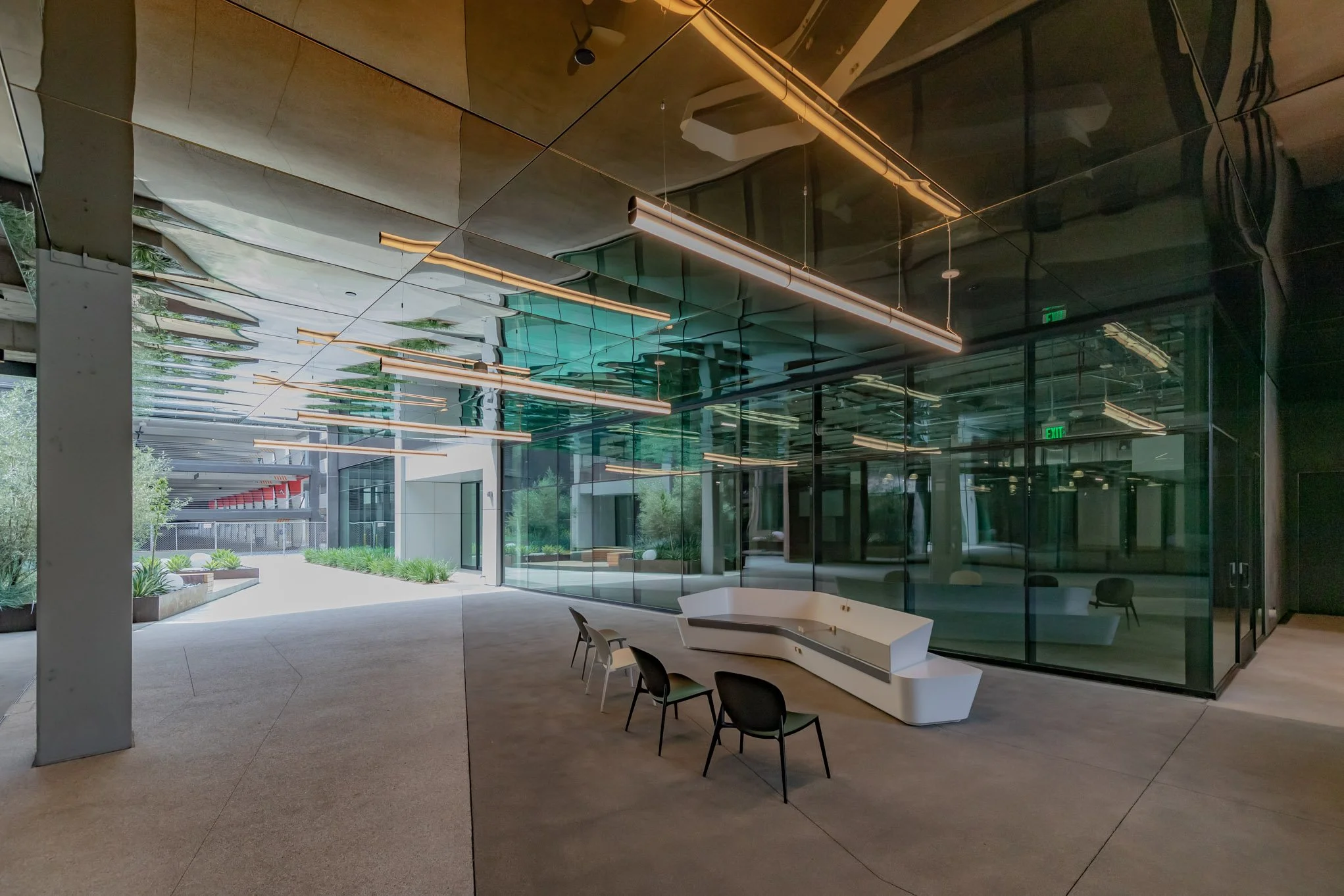 Modern building interior with reflective glass walls, an abstract white seating area, and three black chairs, illuminated by linear ceiling lights. Outside visible are trees and a pathway.