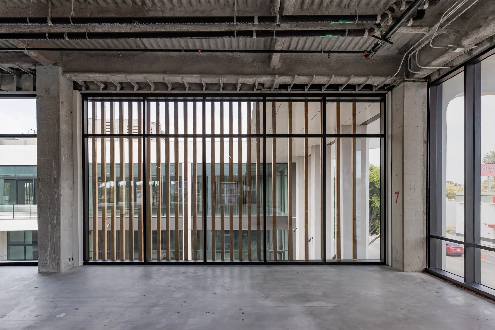 Interior of a building under construction with unfinished concrete floors, exposed ceiling with pipes and wiring, and large glass windows with vertical wooden slats in the foreground.