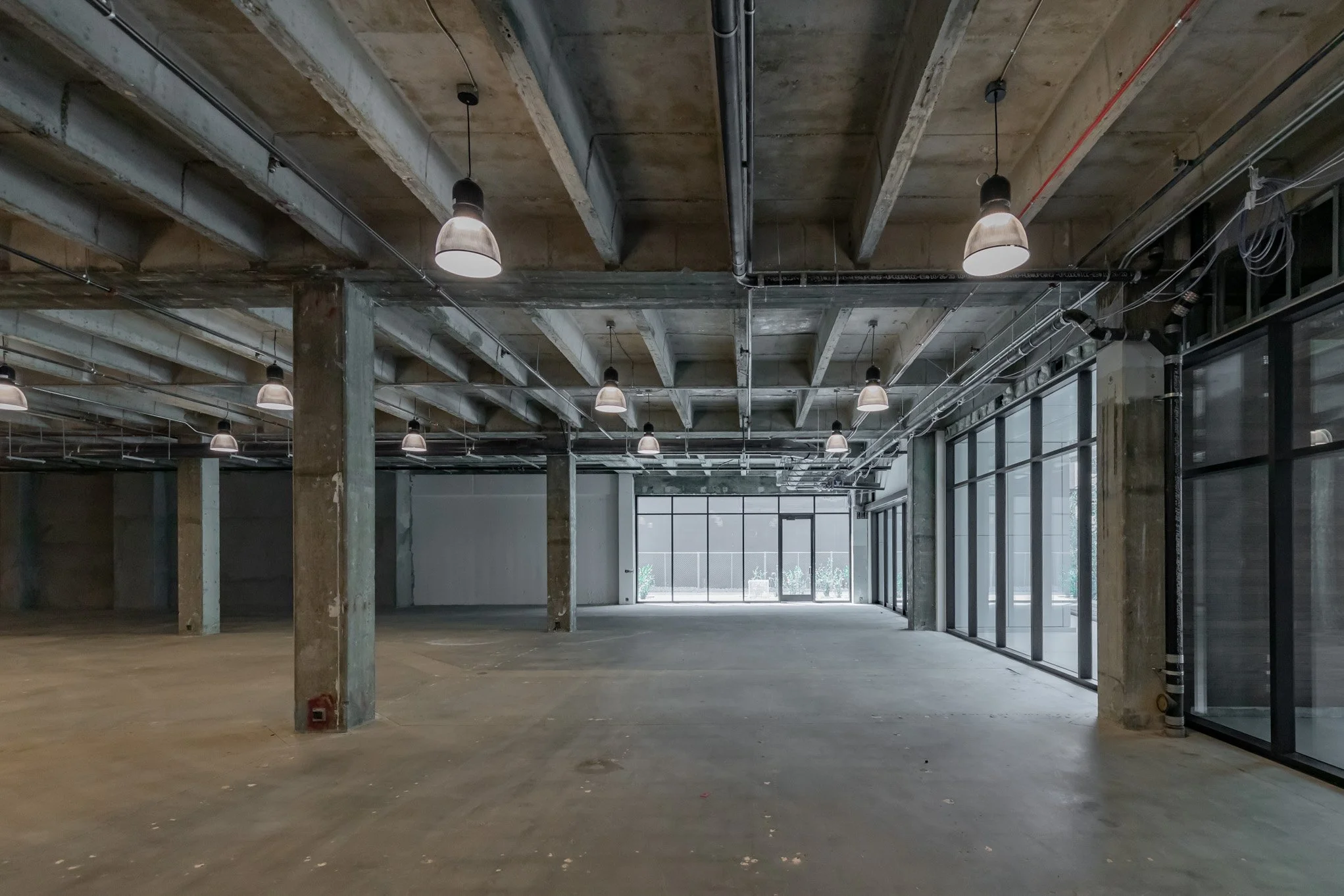 Empty industrial-style commercial space with concrete floors, exposed ceiling beams, black pendant lights, and floor-to-ceiling windows
