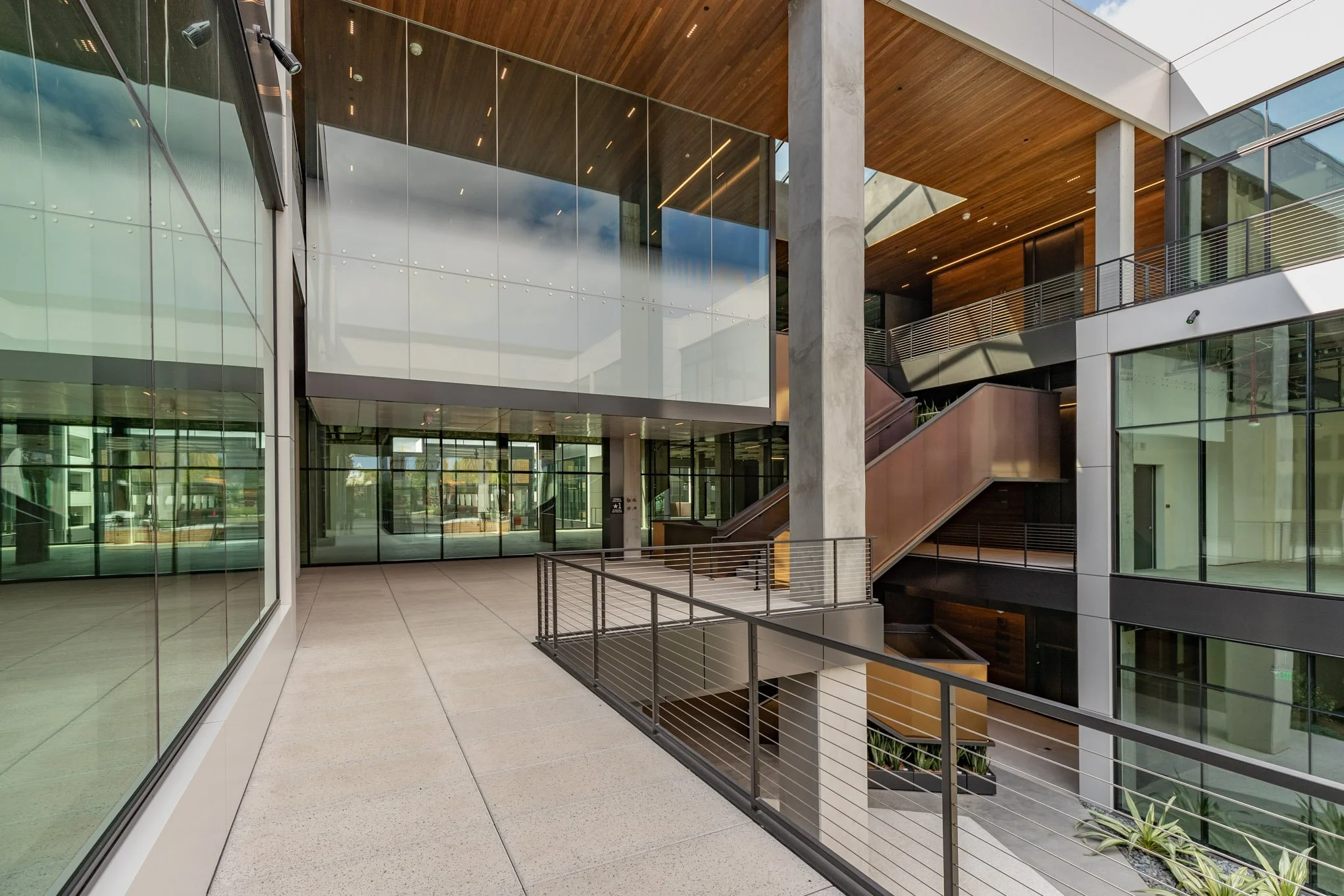 Modern building interior with glass walls, concrete columns, wooden ceiling, and metal railings, featuring multiple levels and stairs.