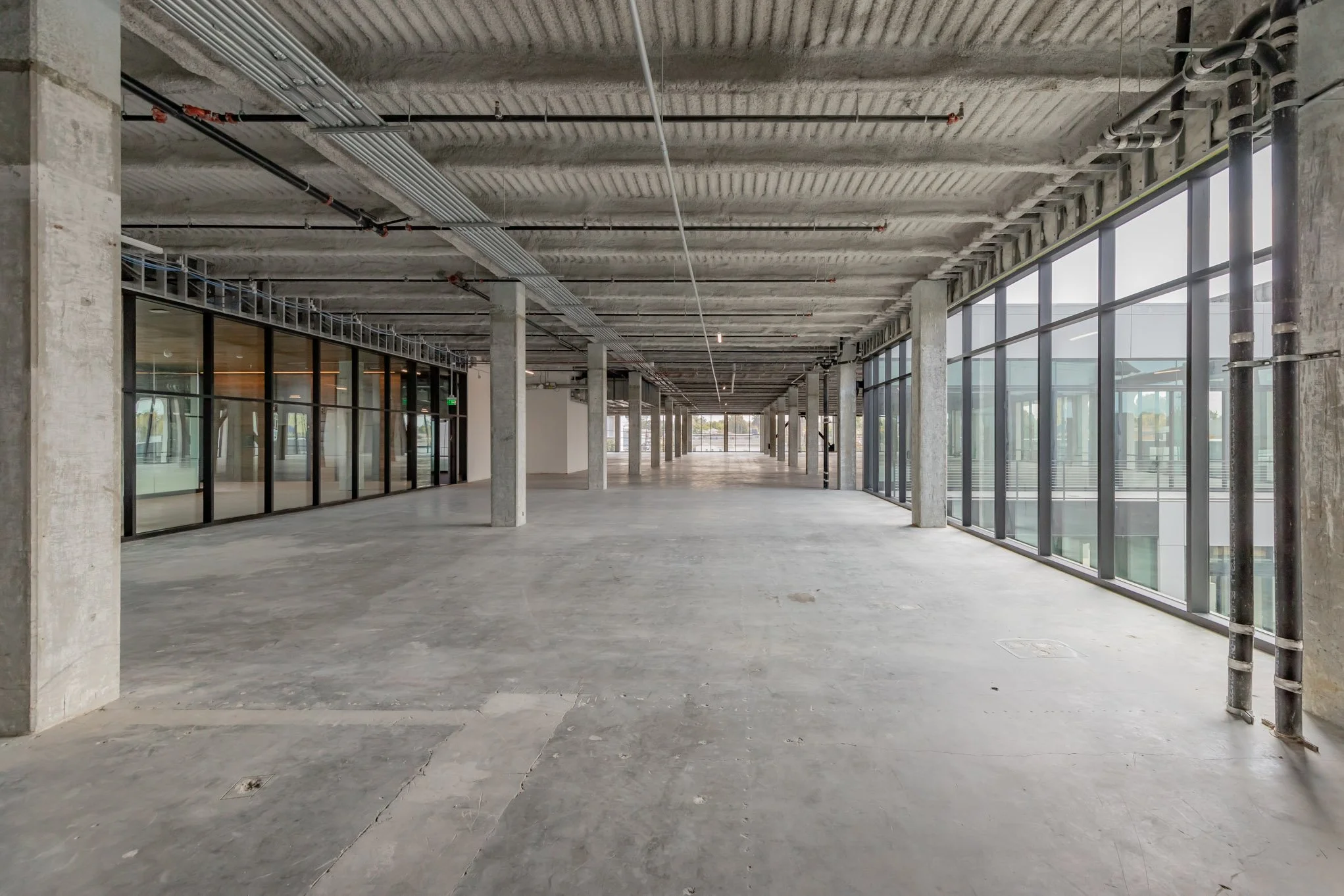 Empty office space under construction with large glass windows, concrete floors, and exposed ceiling piping.