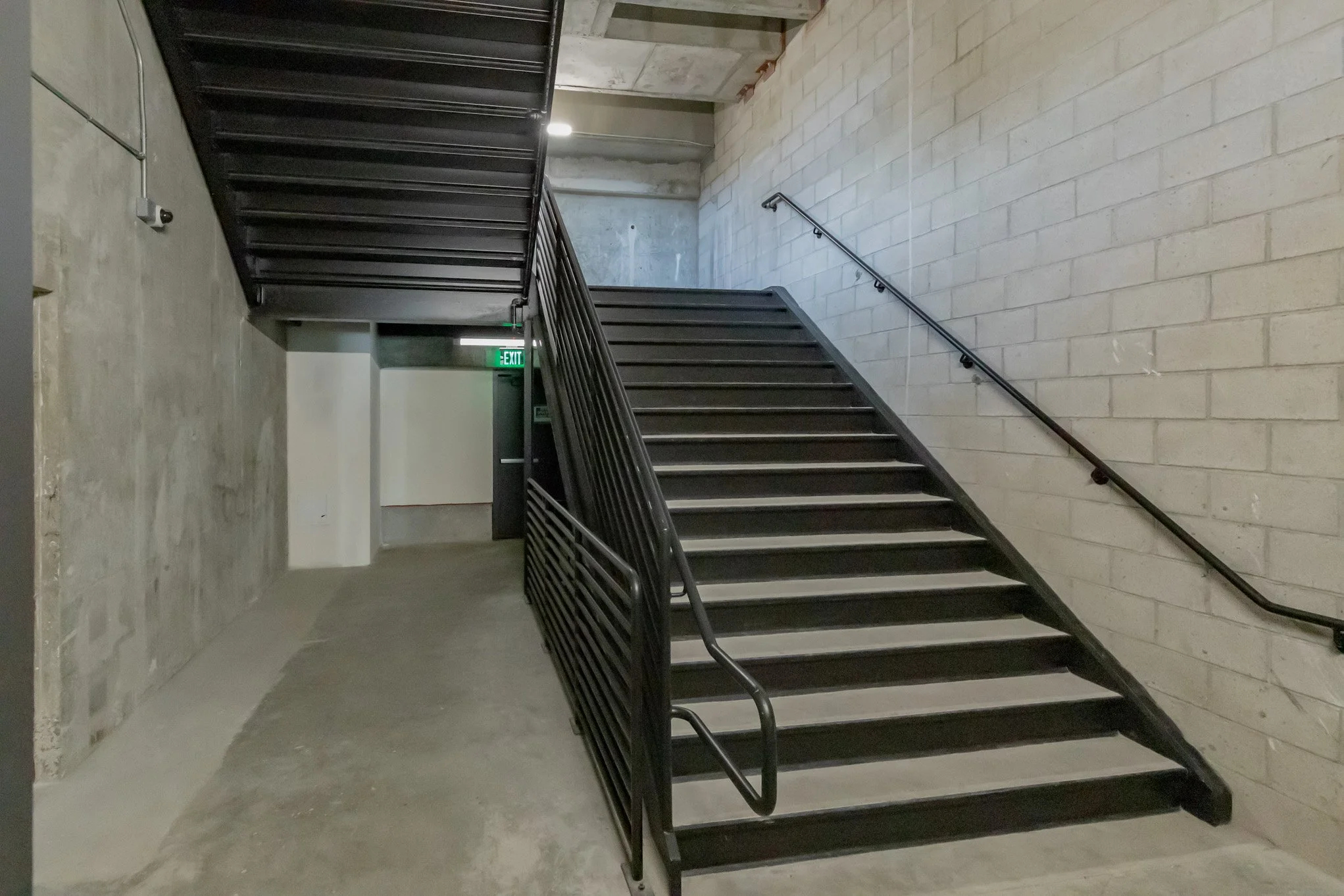 Interior view of a concrete building with black metal stairs and handrails, beige brick wall on the right, and a sign above exit in the background.