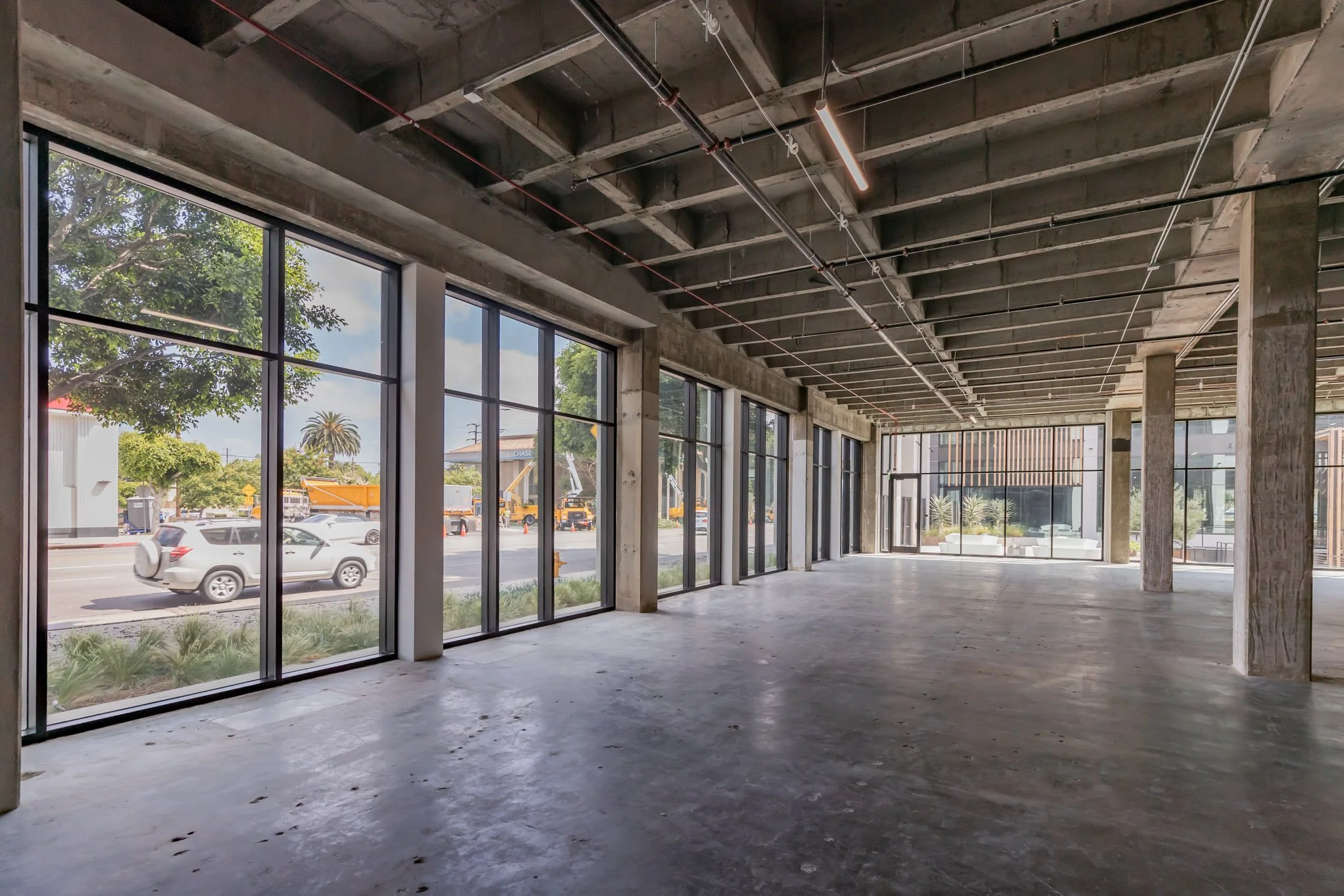 Interior of an unfinished commercial space with large floor-to-ceiling windows, exposed ceiling with pipes and lighting, and empty concrete floor.
