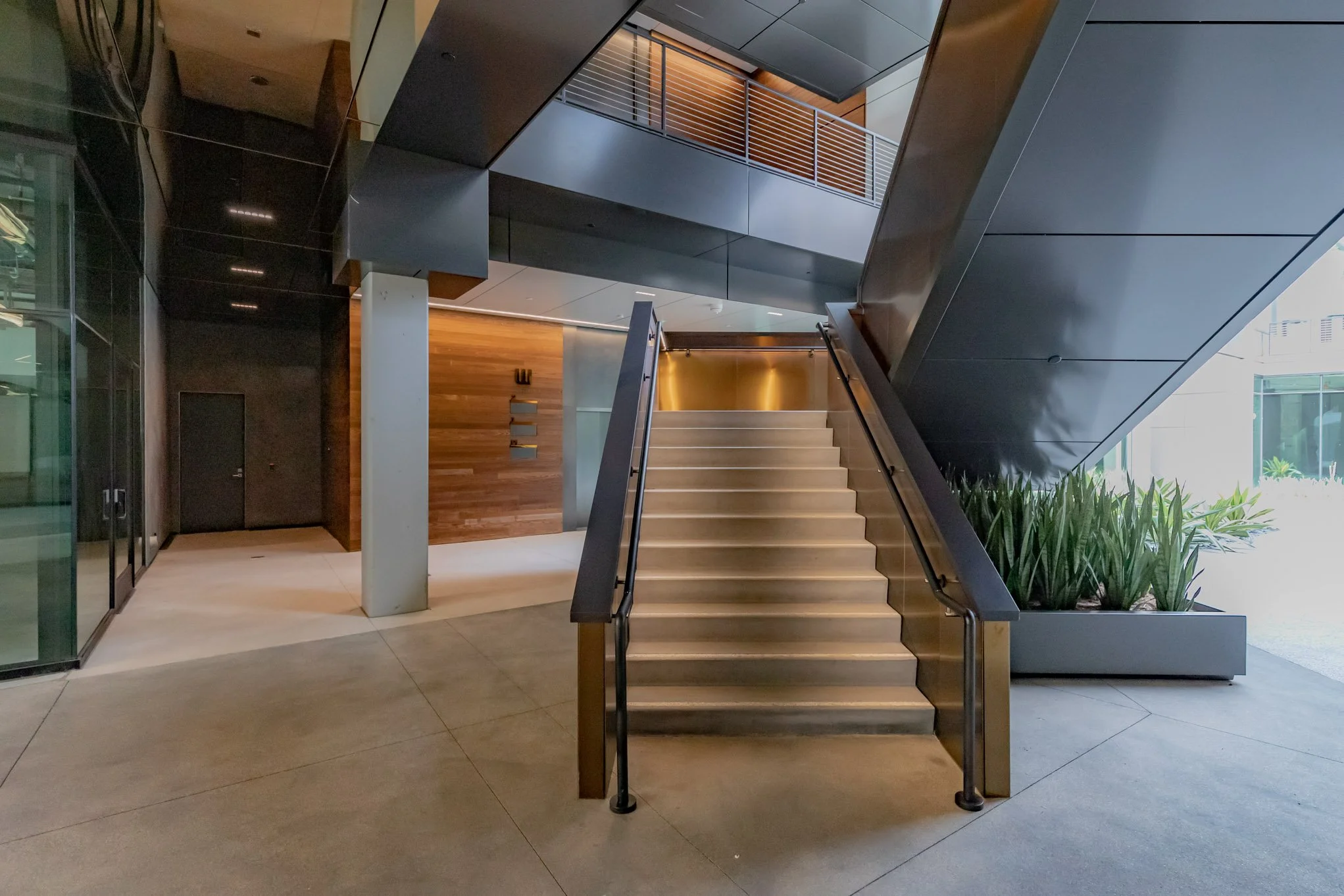 Modern building interior with a staircase, plants, glass walls, and wood paneling.