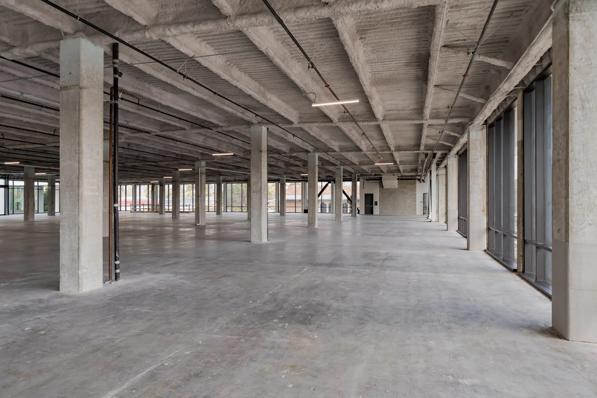 An empty, spacious interior of a commercial building under construction with concrete pillars, unfinished floors, and large windows along one side.
