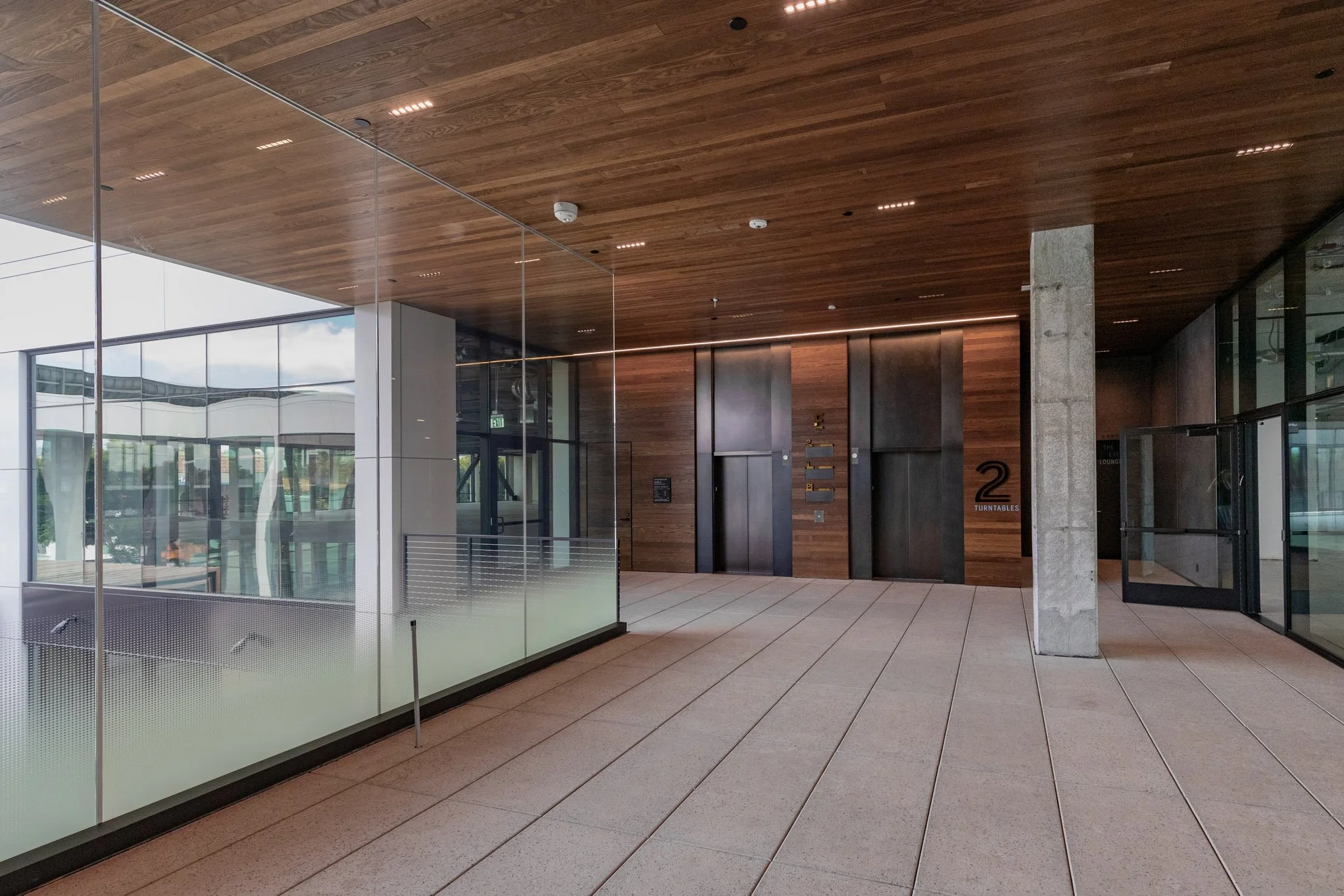 Modern lobby area with wooden ceiling, glass walls, and two elevators with dark doors.