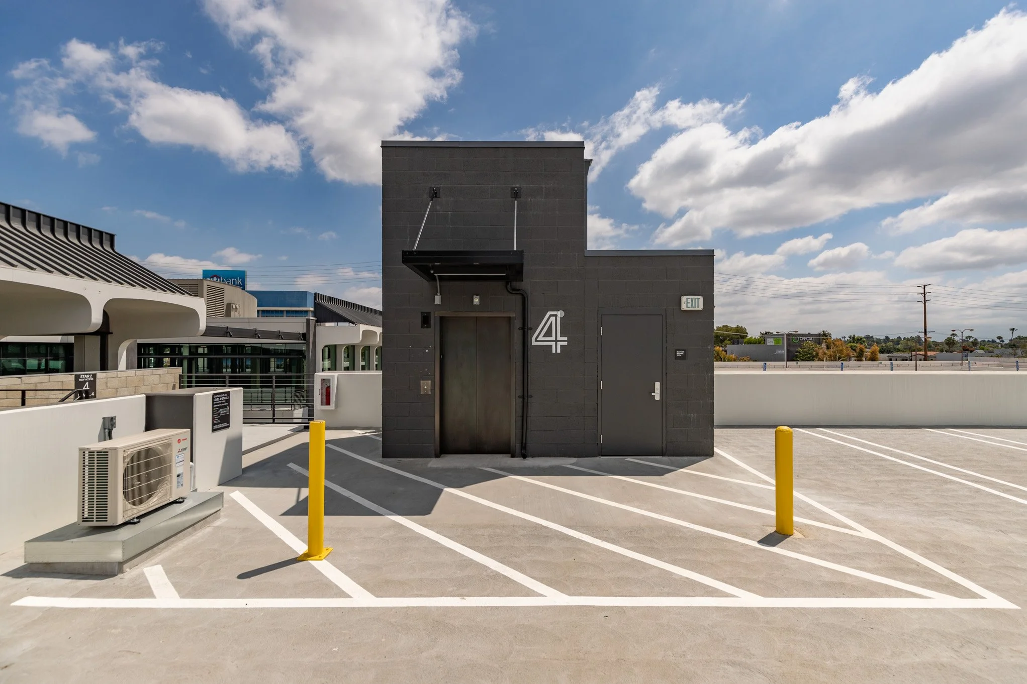 Empty parking lot with yellow parking barriers in front of a black building with an elevator, door, and exit sign under a partly cloudy sky.